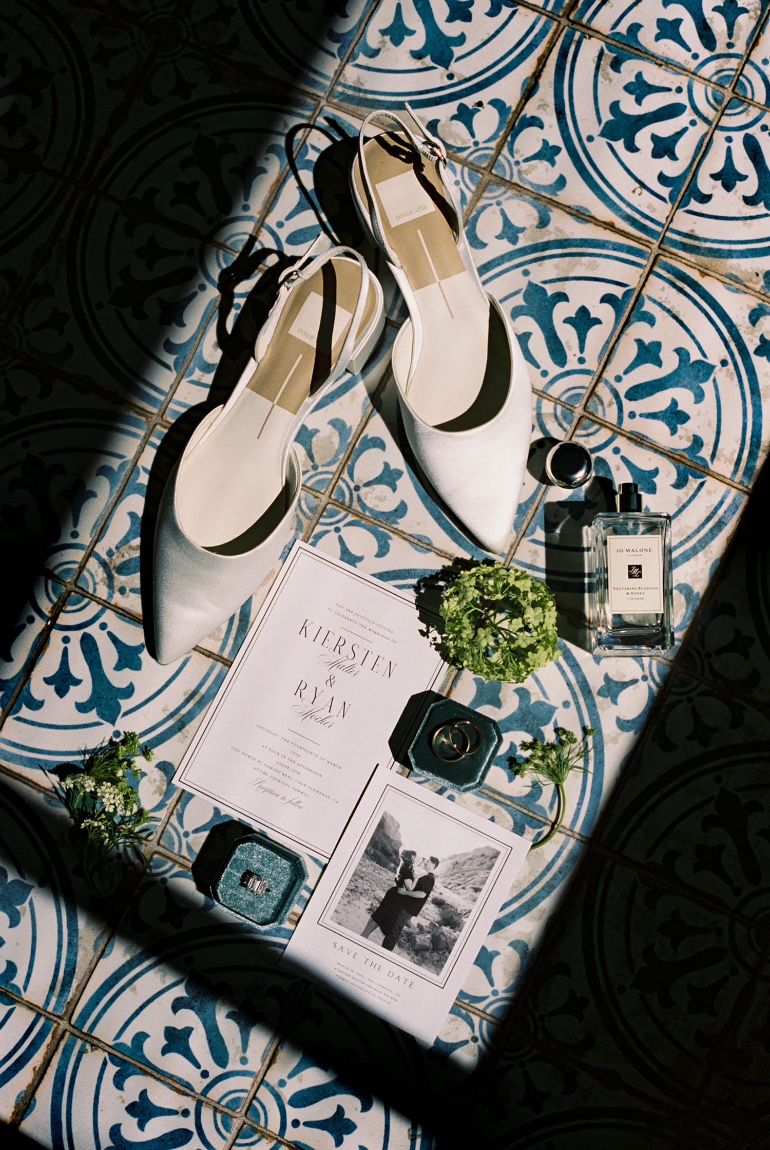 Elegant wedding flat lay with white heels, Jo Malone perfume, ring boxes, floral greenery, and invitations on blue tile.
