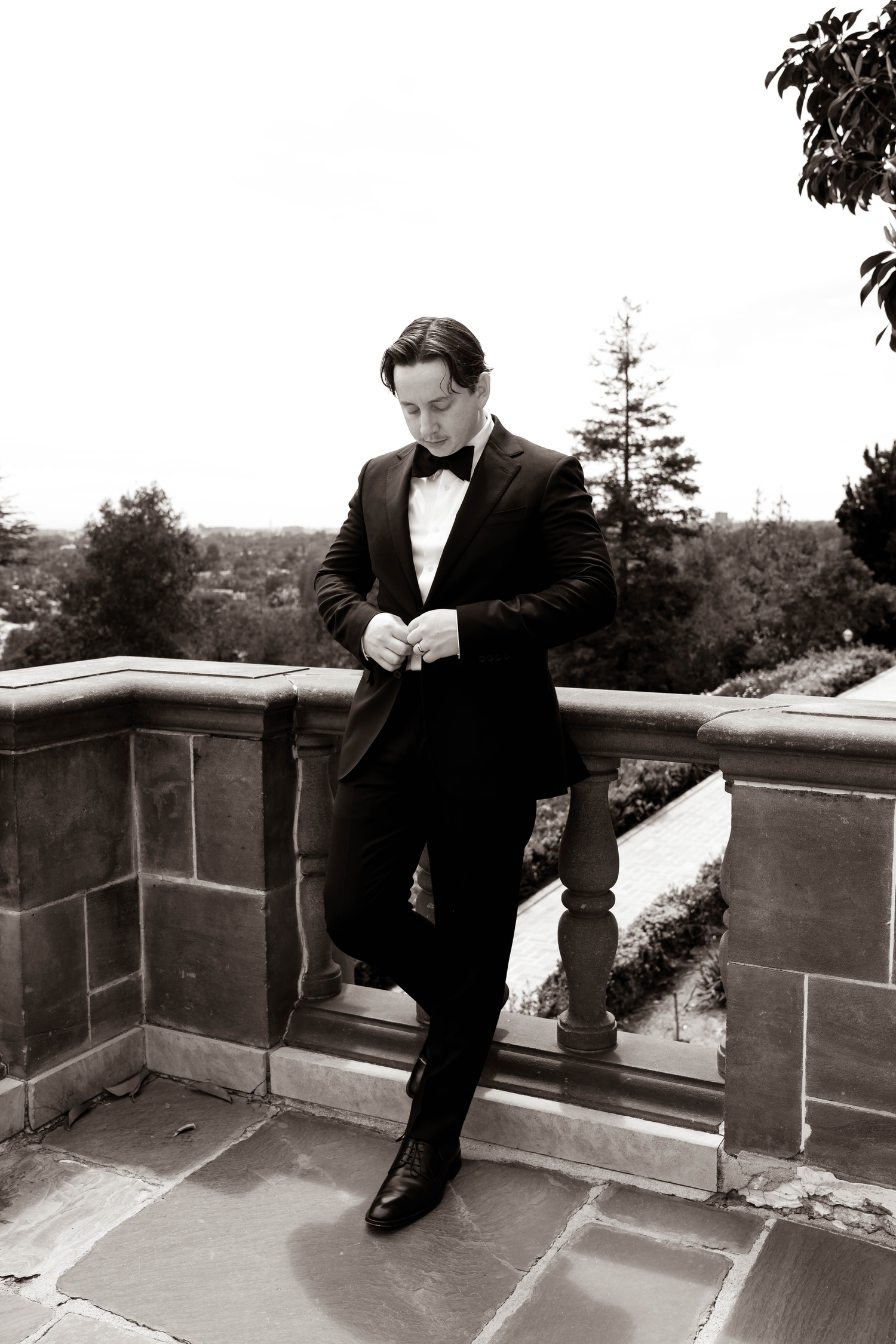 A dapper groom in a black tuxedo and bow tie adjusts his jacket on a stone terrace in this elegant black and white photo.
