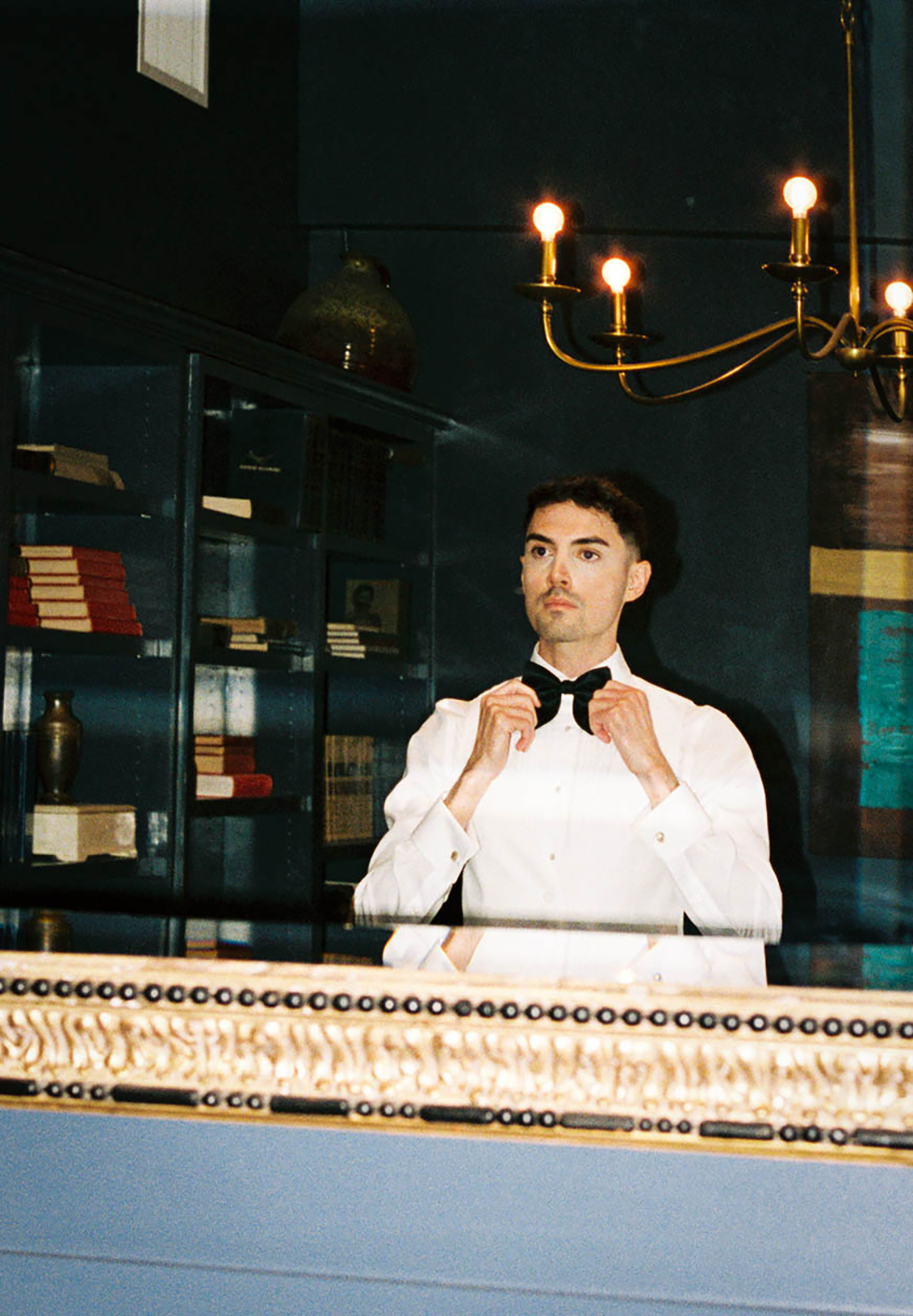 Man in white dress shirt adjusting black bow tie in elegant room with brass chandelier and display cases.