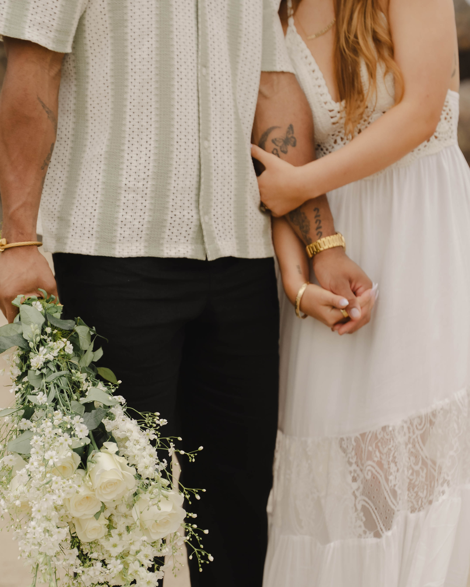 Couple holding hands at wedding, woman in white lace dress with bouquet of white flowers and greenery.