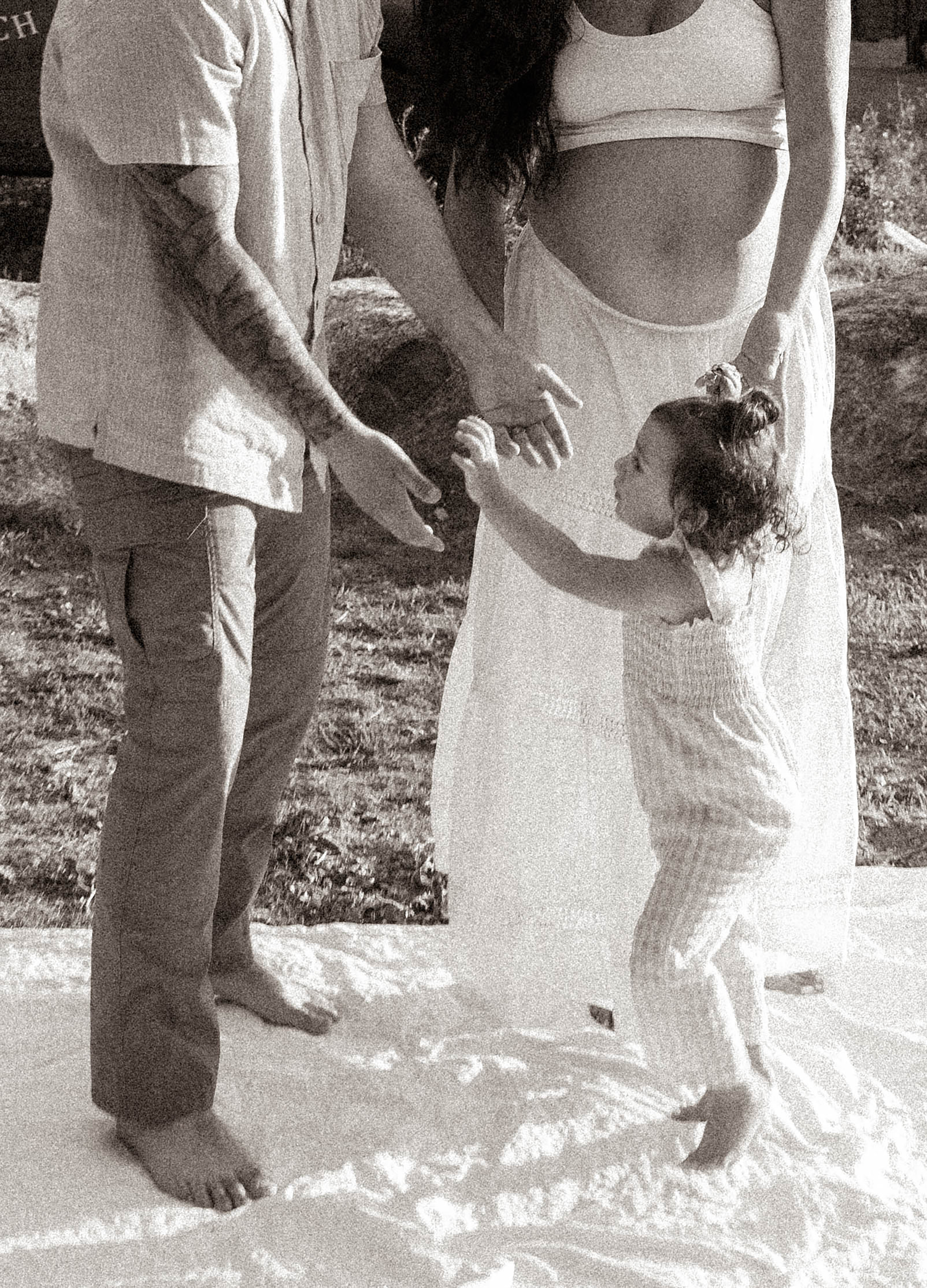 Black and white maternity photo of pregnant woman, tattooed man, and toddler girl holding hands outdoors.