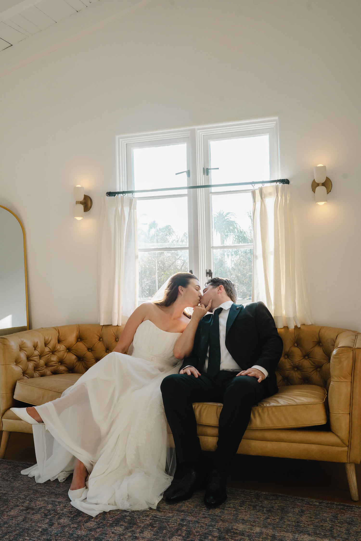 Bride and groom sharing a kiss while seated on a tufted leather sofa in an elegant wedding venue.
