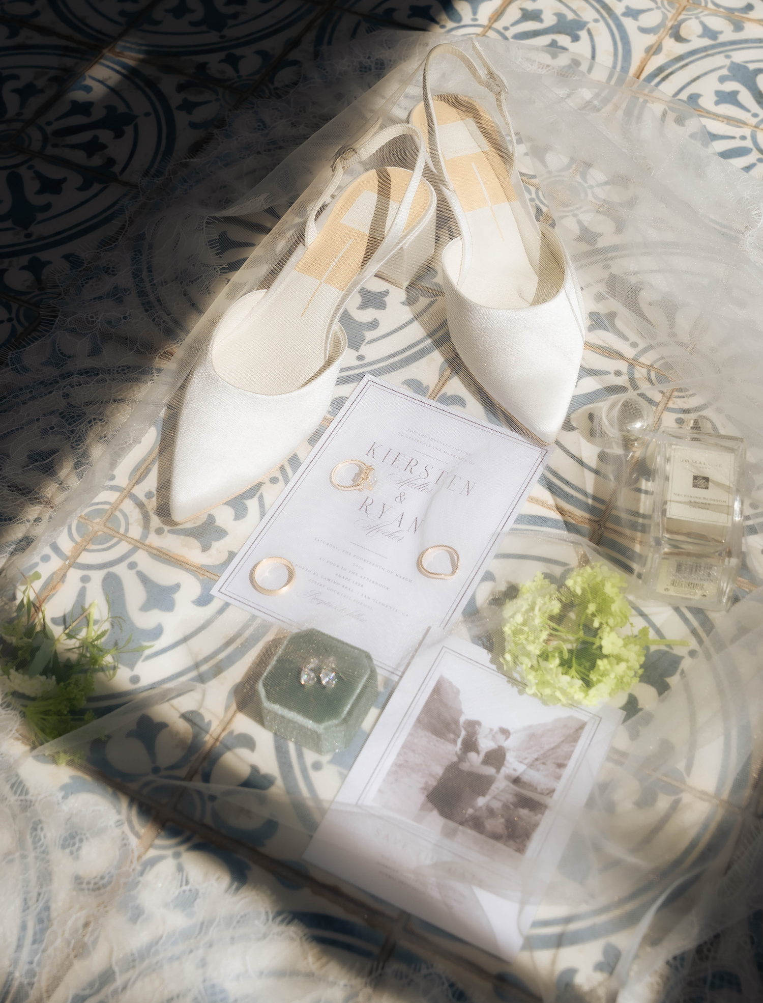 Elegant bridal flat lay featuring white satin heels, gold wedding rings, stud earrings, perfume bottle, and wedding invitation on tile.