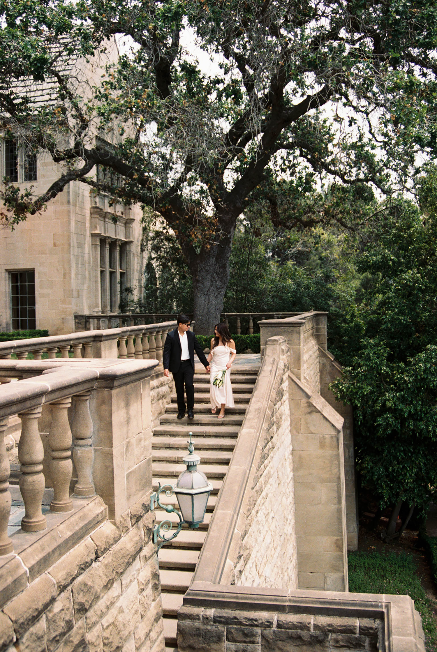 Couple in wedding attire walks down elegant stone staircase beside historic building surrounded by lush green trees.