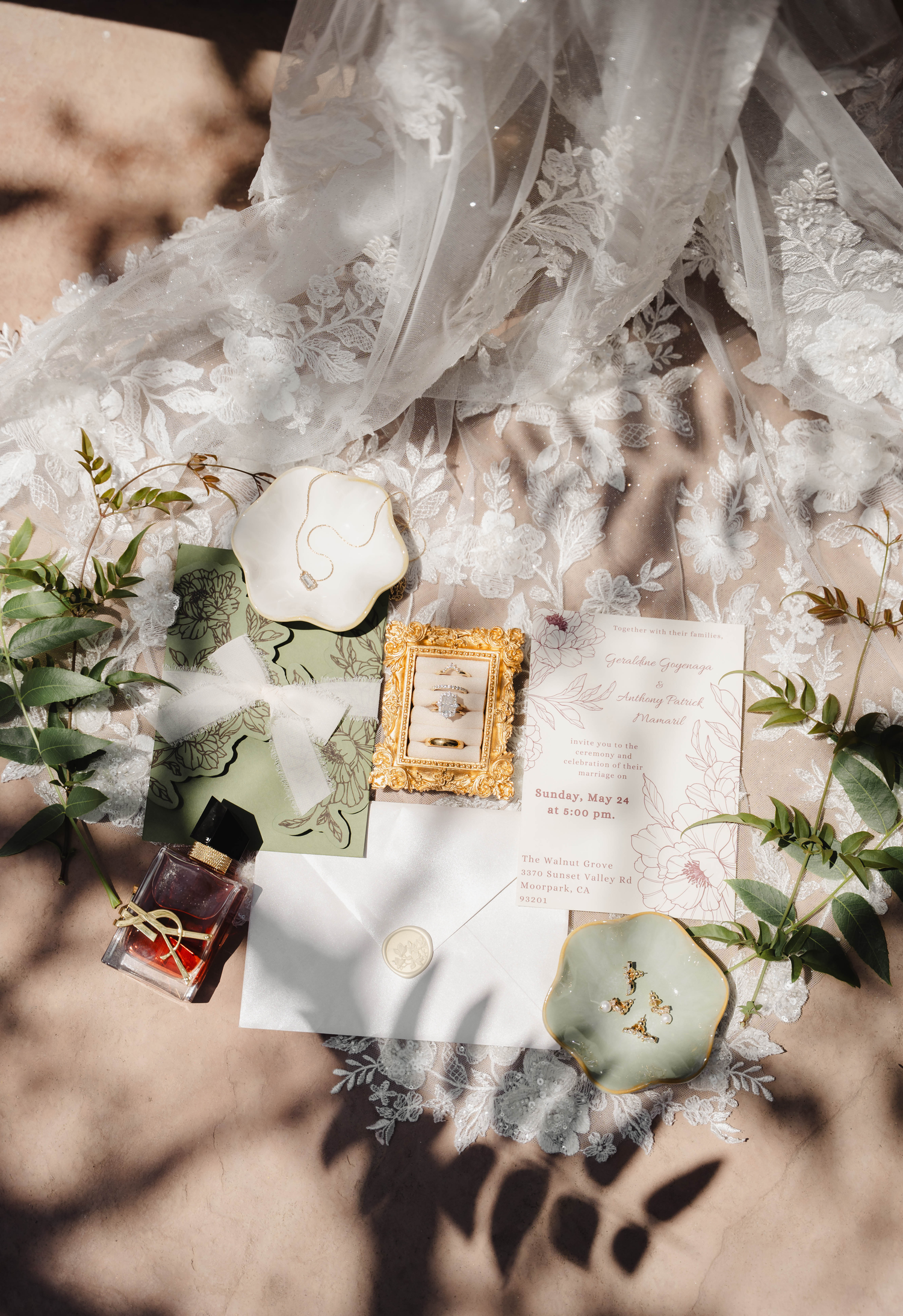 Elegant wedding flat lay with lace bridal gown, green envelopes, gold frame, rings, perfume, and floral greenery on neutral background.
