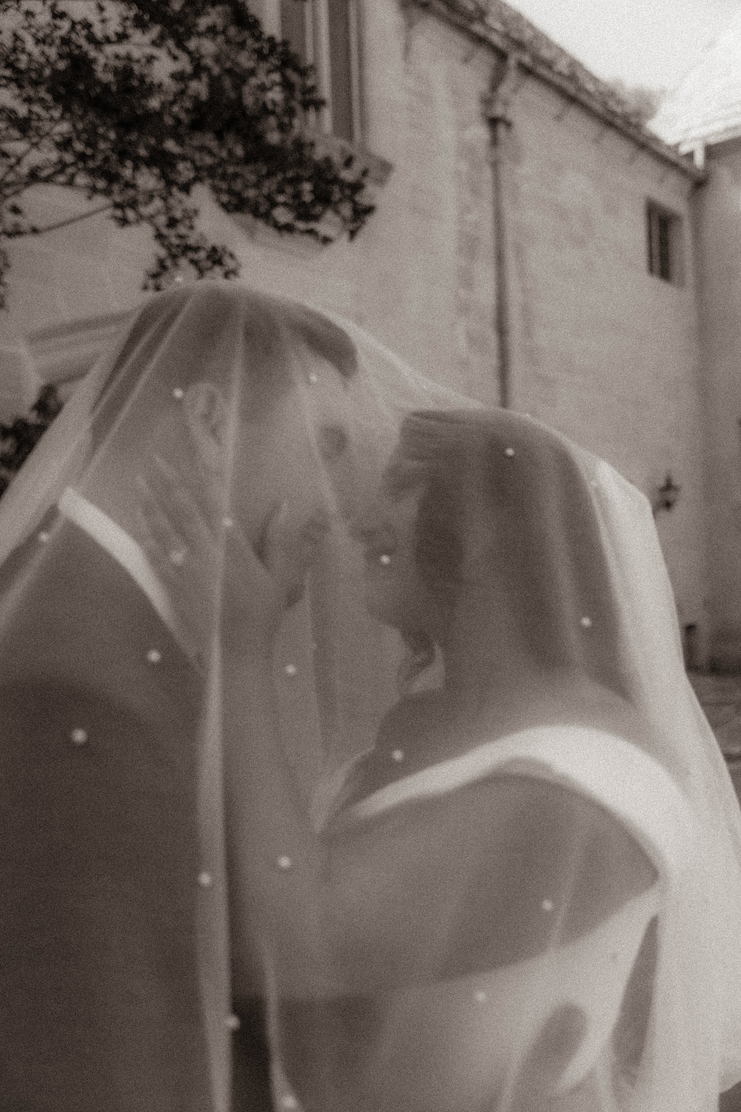 Romantic black and white photo of newlyweds nearly kissing beneath a dotted wedding veil outdoors.