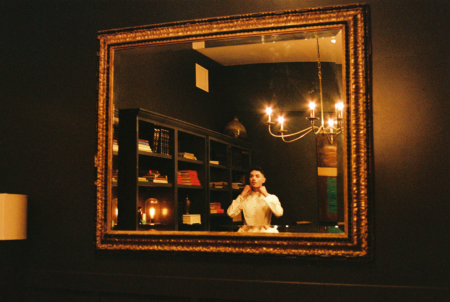 Person photographed in mirror reflection with ornate gold frame, chandelier, and dark wood shelving visible in background.