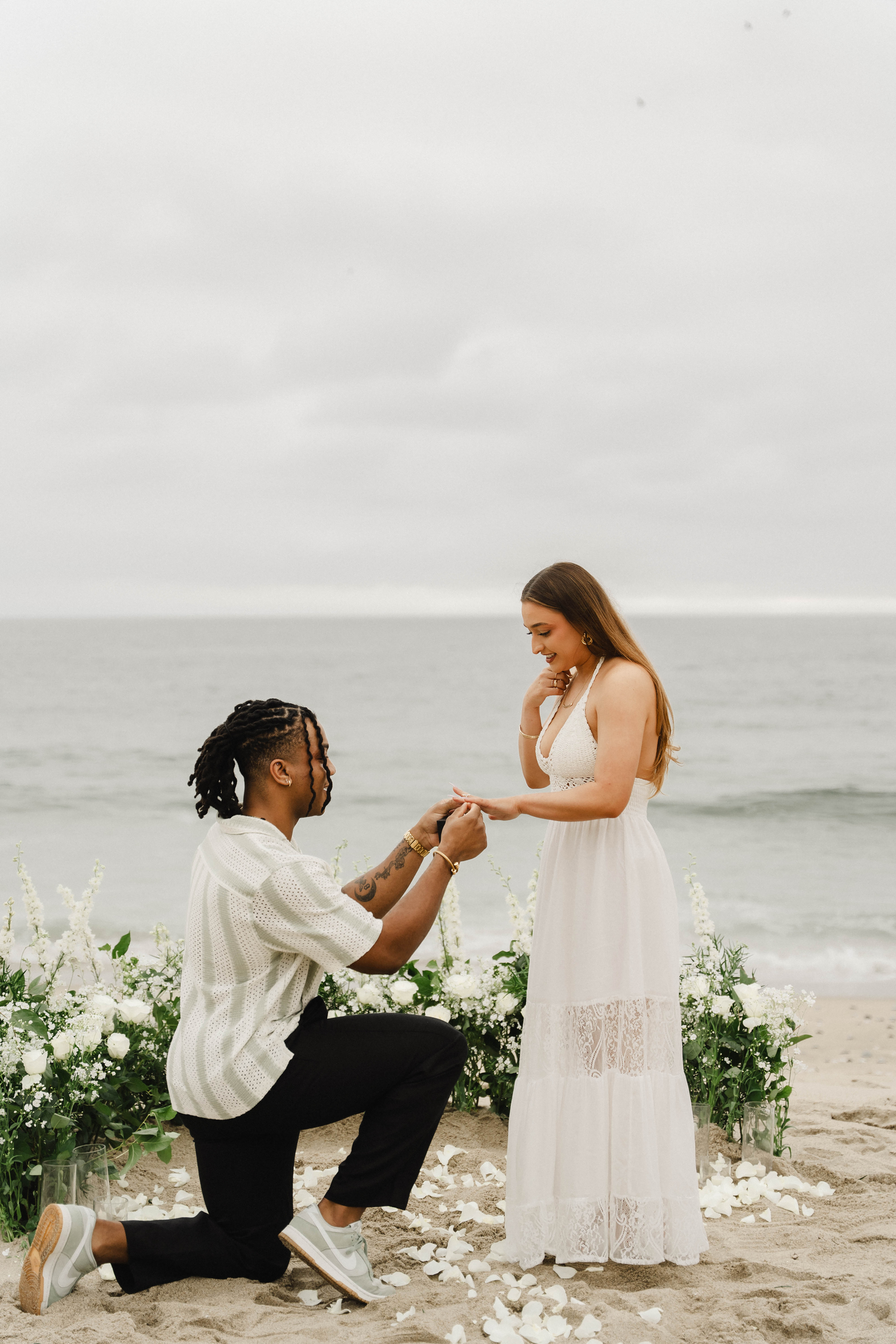 Man proposes on one knee to woman in white dress on sandy beach with floral arrangements and ocean backdrop.