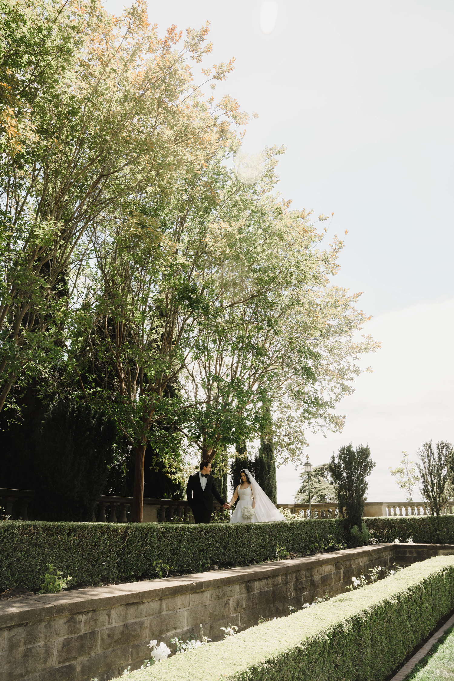 A bride and groom walk hand in hand through a lush formal garden with manicured hedges and tall trees.