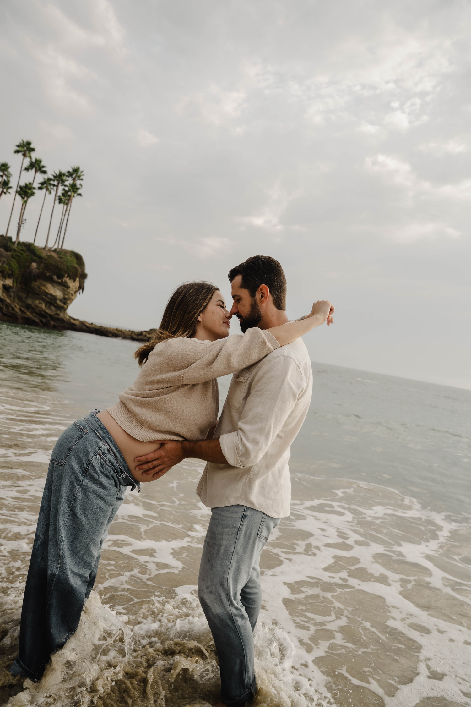 Expecting couple sharing a tender nose-to-nose moment on a rocky beach with palm trees and ocean waves.