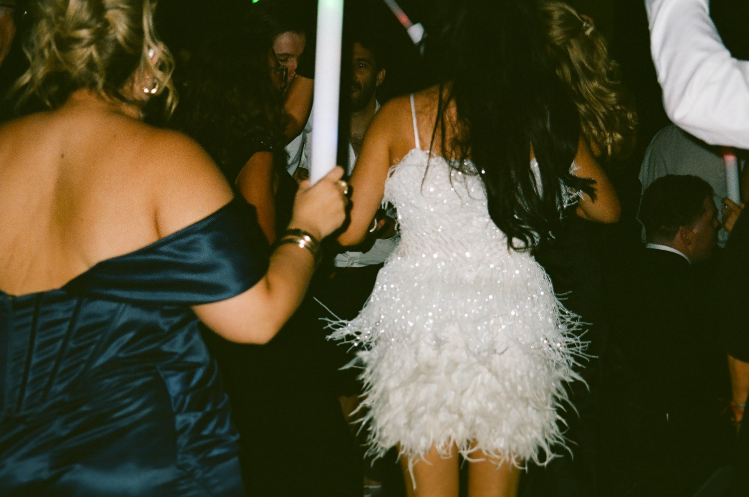 Women dancing at a party, one wearing a white feathered mini dress, another in a blue satin gown, at a nighttime event.