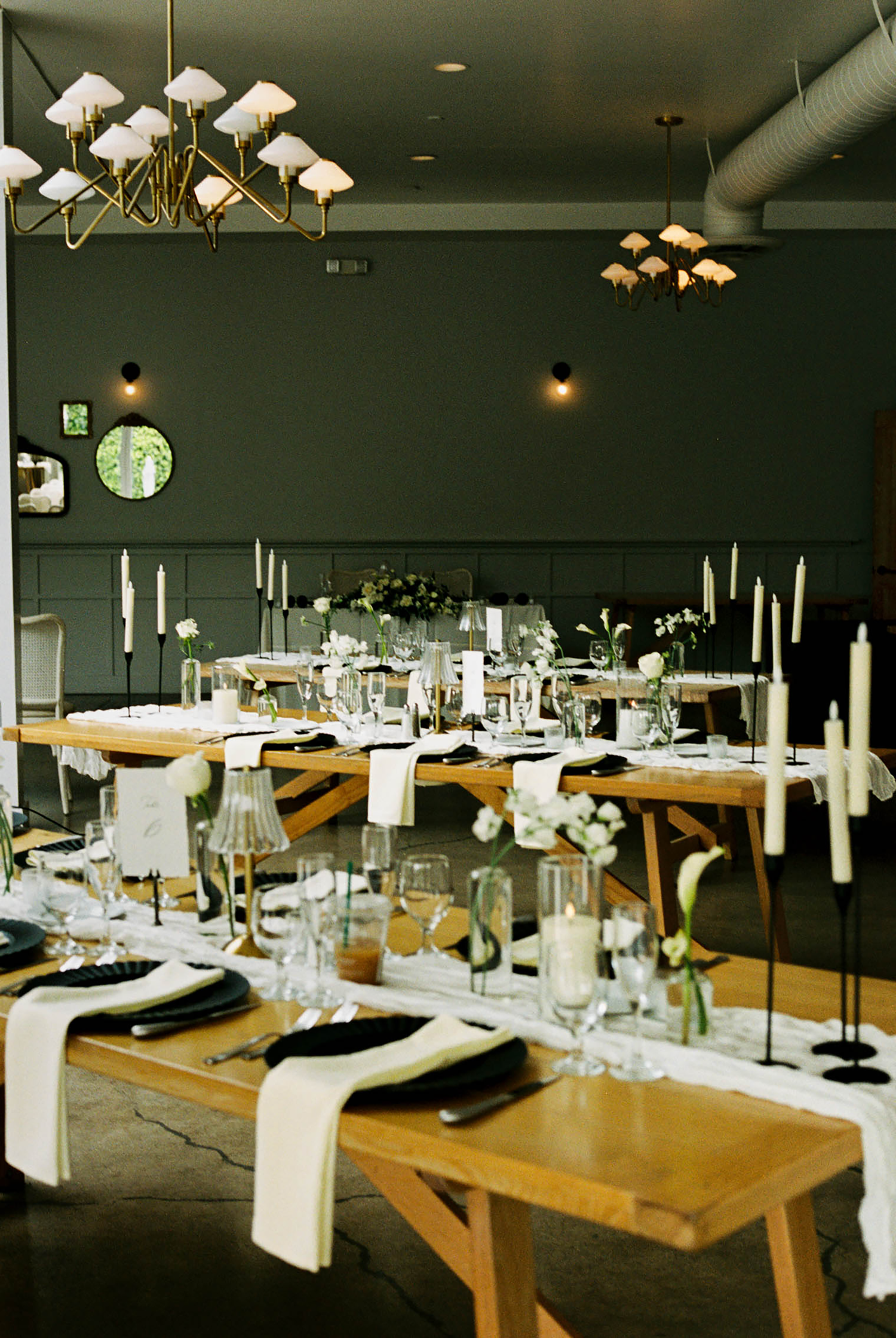 Elegant wedding reception with wooden tables, black plates, white tapered candles, and floral centerpieces in a dark-walled venue.