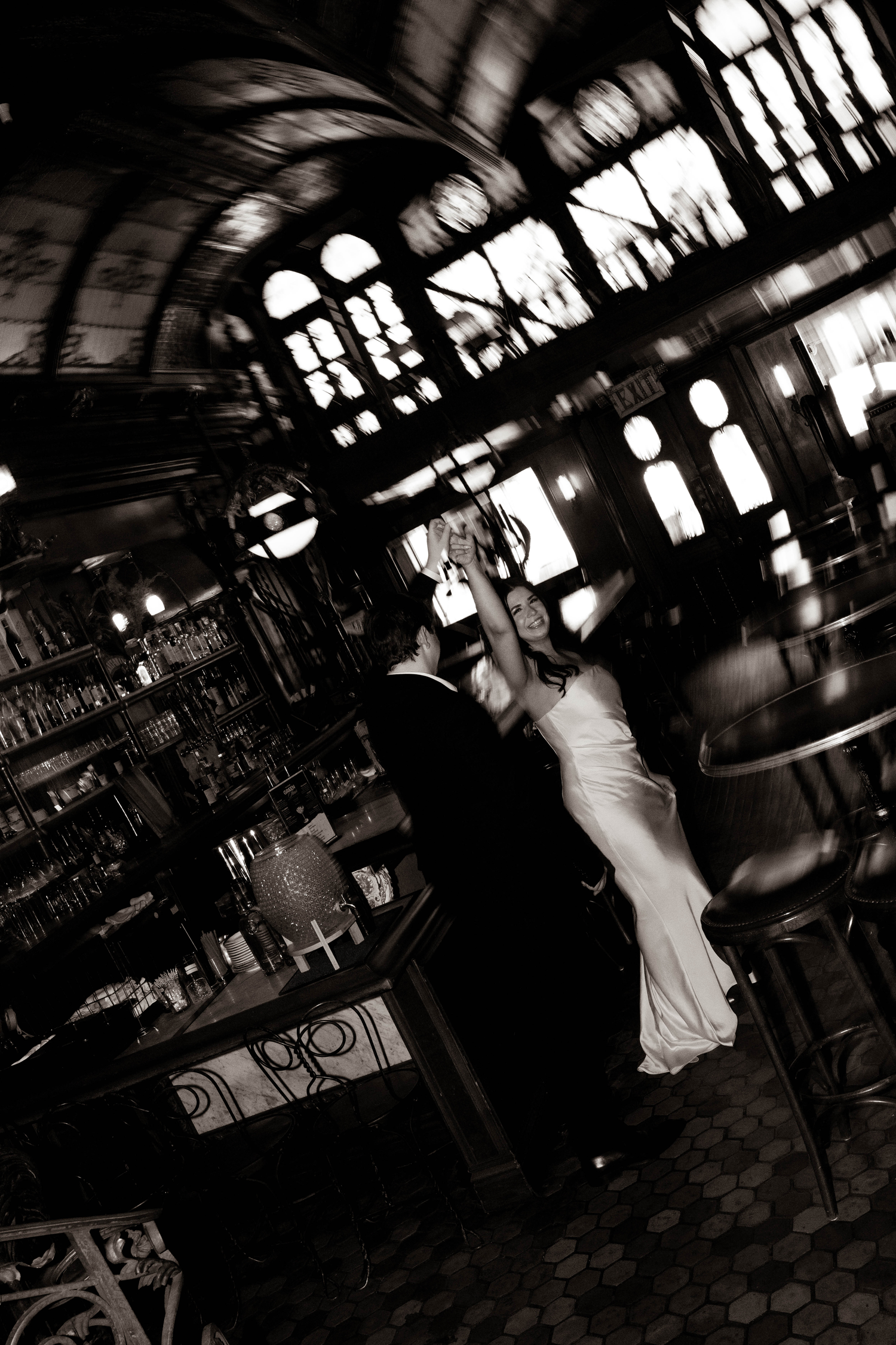 Black and white photo of a bride and groom dancing in an elegant vintage bar with ornate windows and warm lighting.