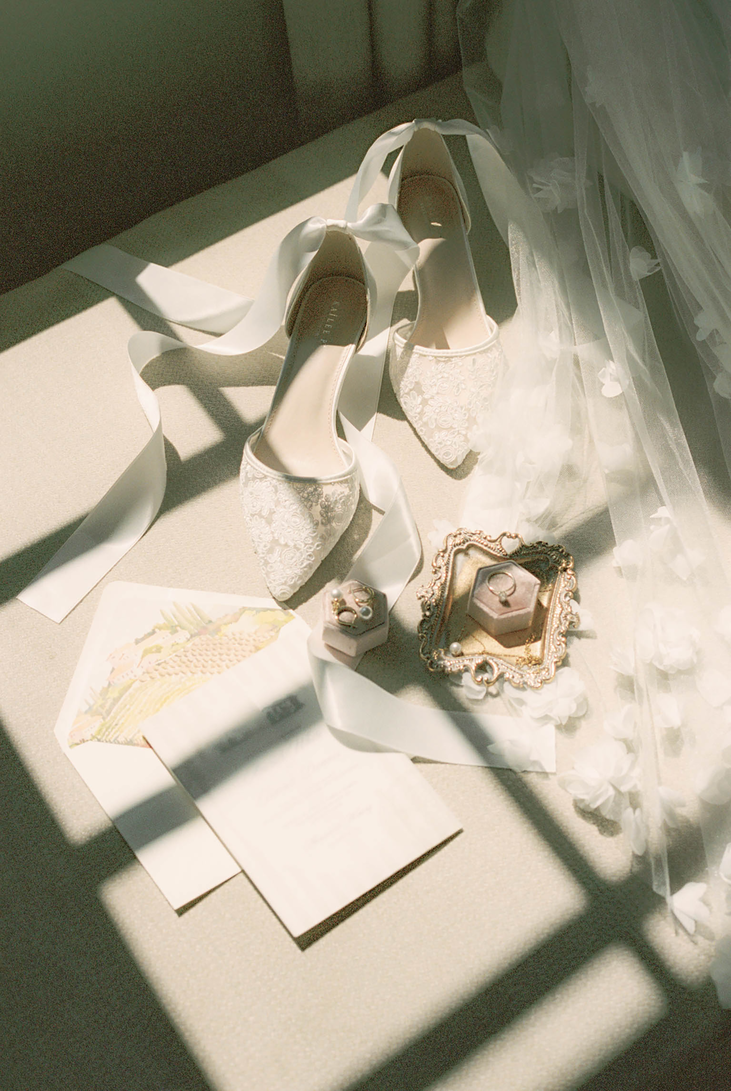 White lace bridal heels with ribbon ties beside a ring box and veil in soft natural window light.