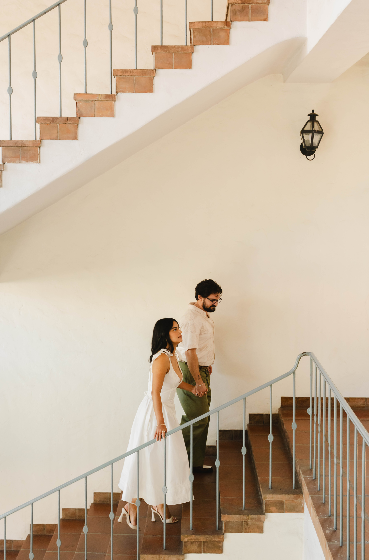 A couple walks up a sunlit staircase, woman in white dress and man in green pants holding hands.