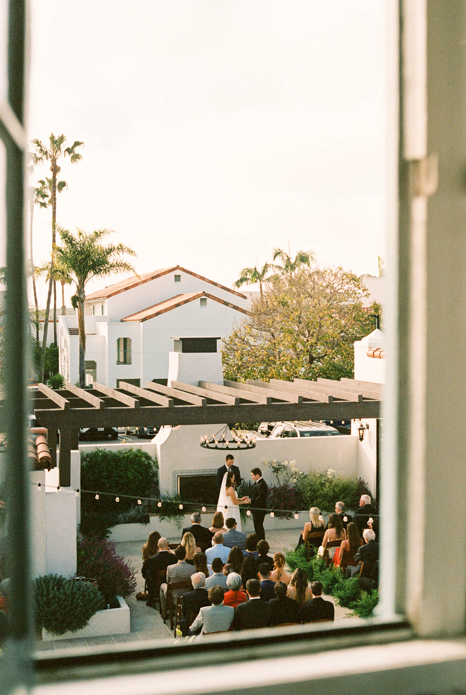 Outdoor wedding ceremony viewed from above through a window at a Spanish-style venue with palm trees.