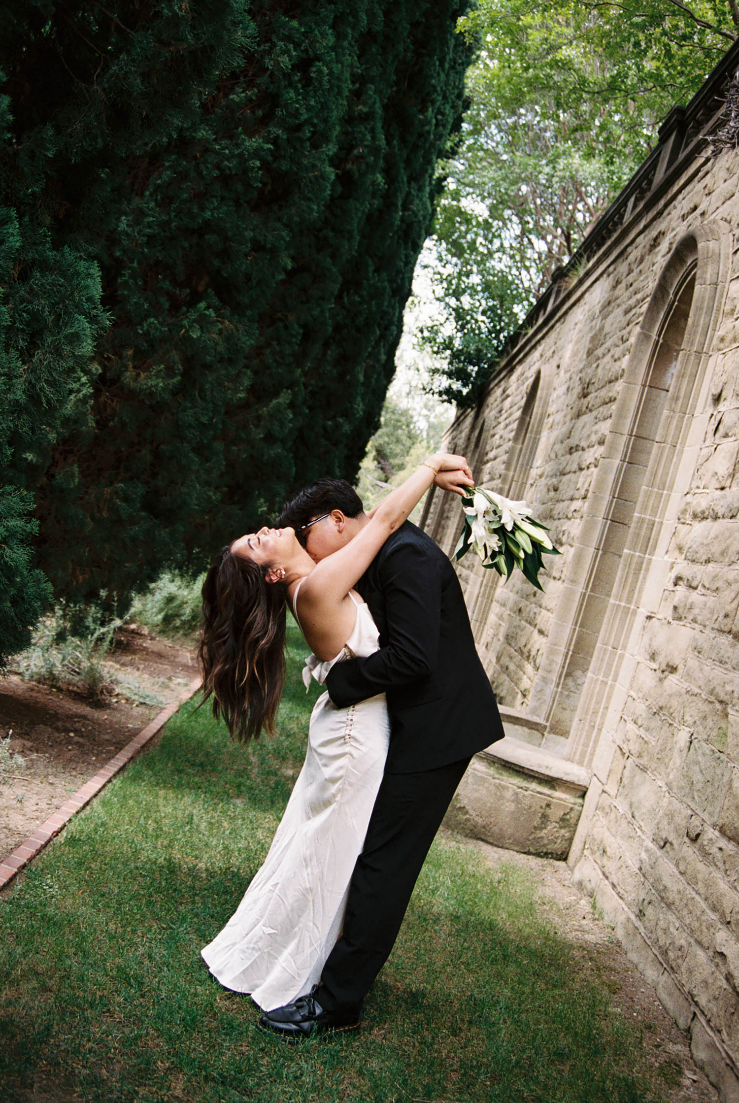 A groom dips his bride in a romantic kiss outdoors beside a stone wall, she holds a white bouquet.