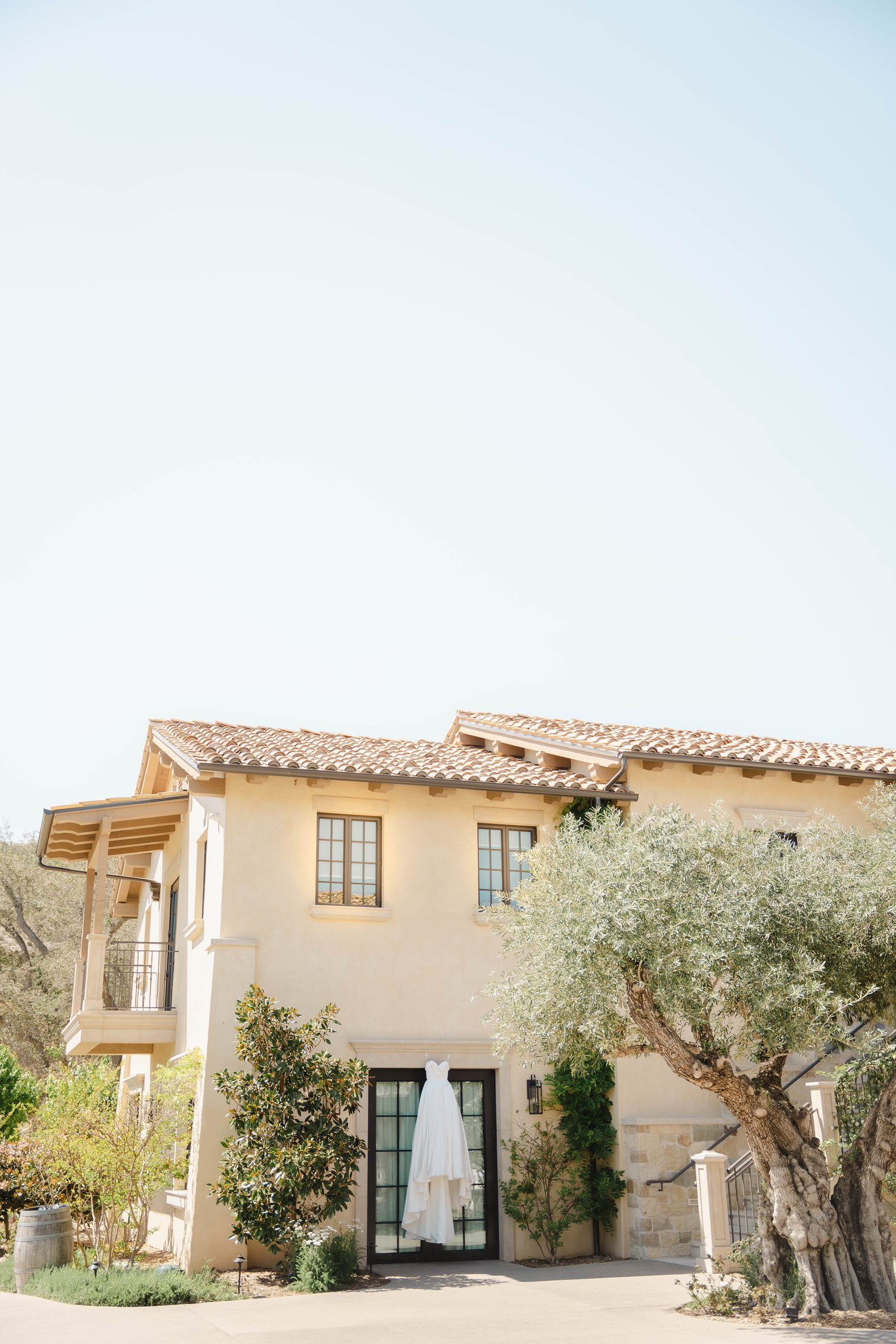 A white wedding dress hangs on a Mediterranean-style villa with terracotta roof and olive tree.