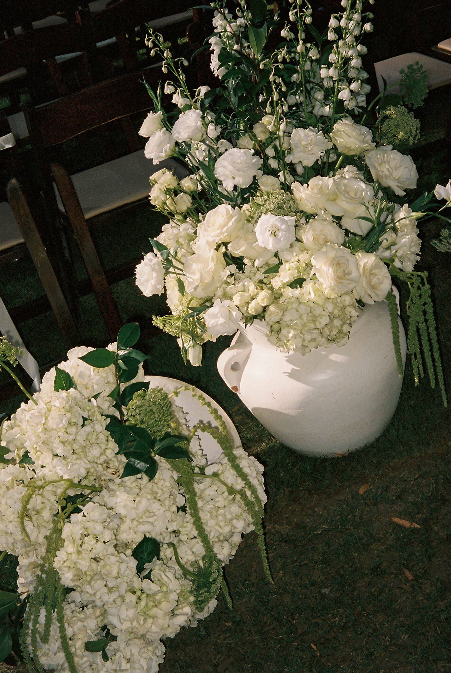 White hydrangeas and roses arranged in a ceramic vase and round wreath for an elegant outdoor wedding ceremony.