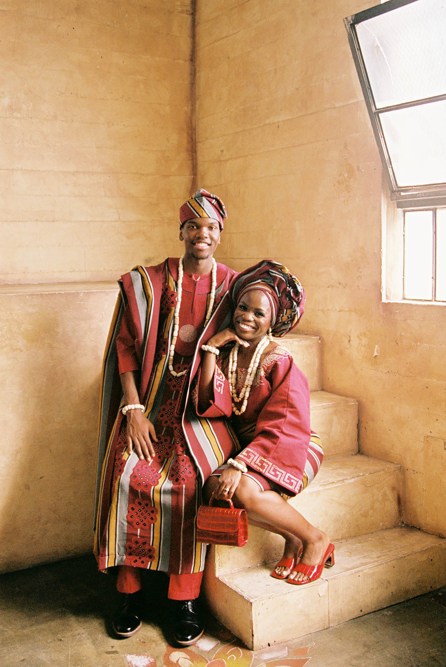 Couple in vibrant traditional African attire with colorful head wraps and beaded jewelry posing on stone steps.