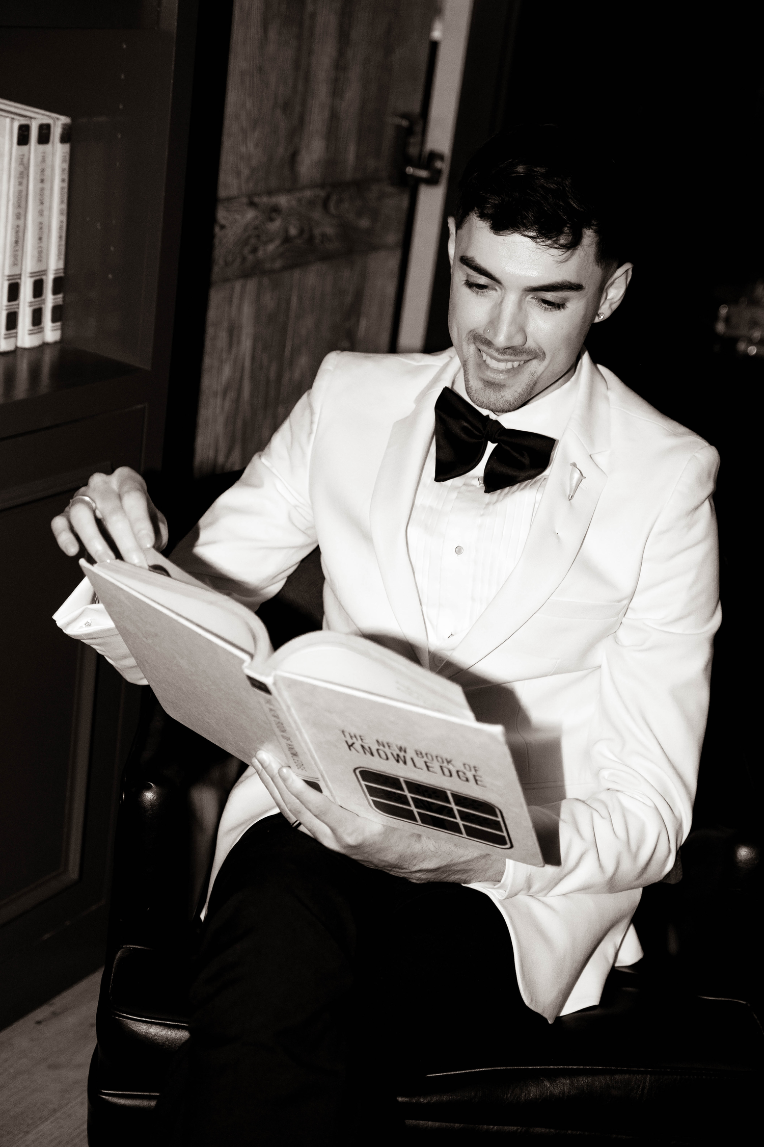 Man in white tuxedo jacket and bow tie smiling while reading a book, black and white photo.