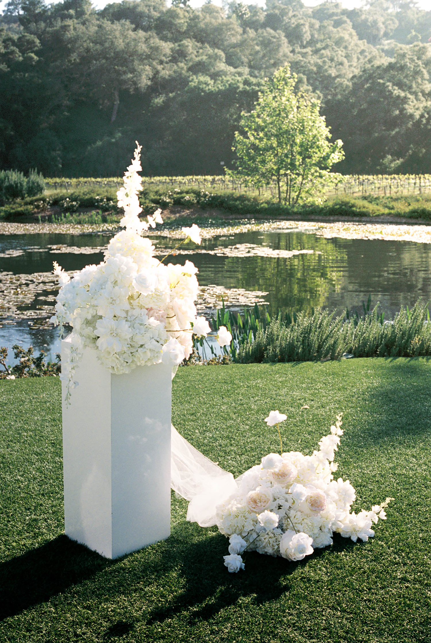 White floral arrangement on pedestal by serene pond at outdoor wedding ceremony.