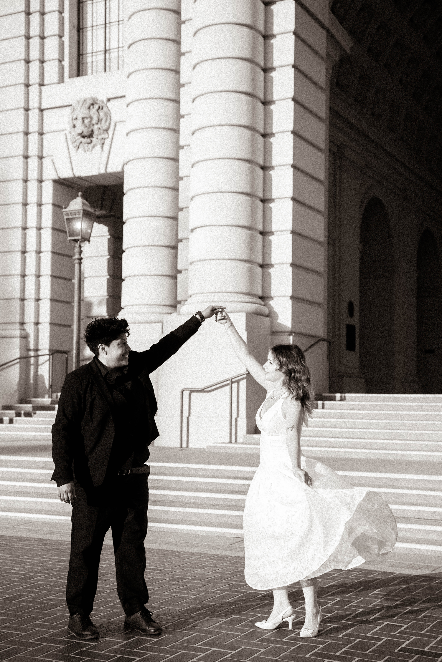 A couple dances in black and white outside a grand building, man in black twirling woman in flowing white dress.