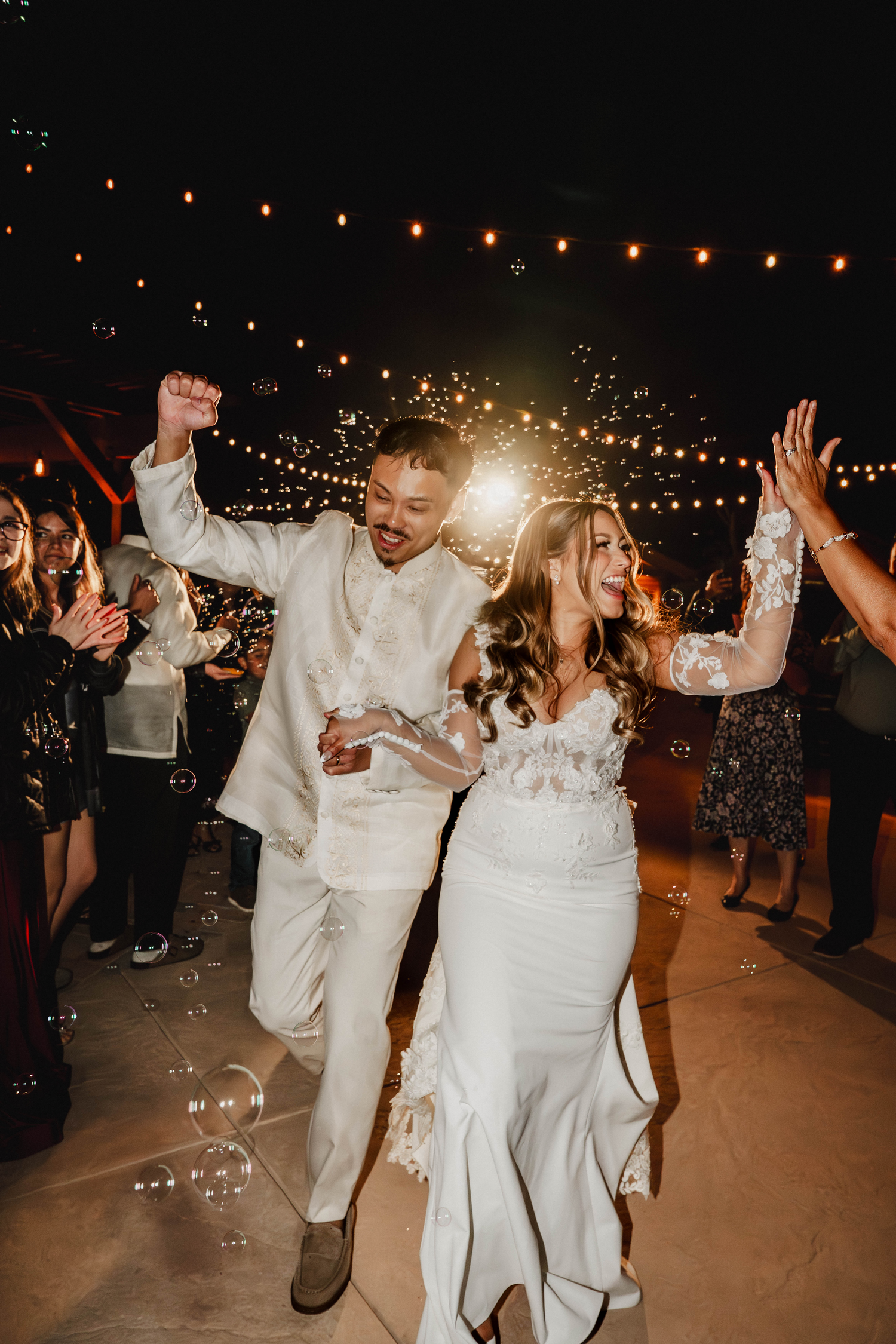 Joyful newlyweds in white celebrate their wedding reception exit with sparklers, bubbles, and cheering guests at night.