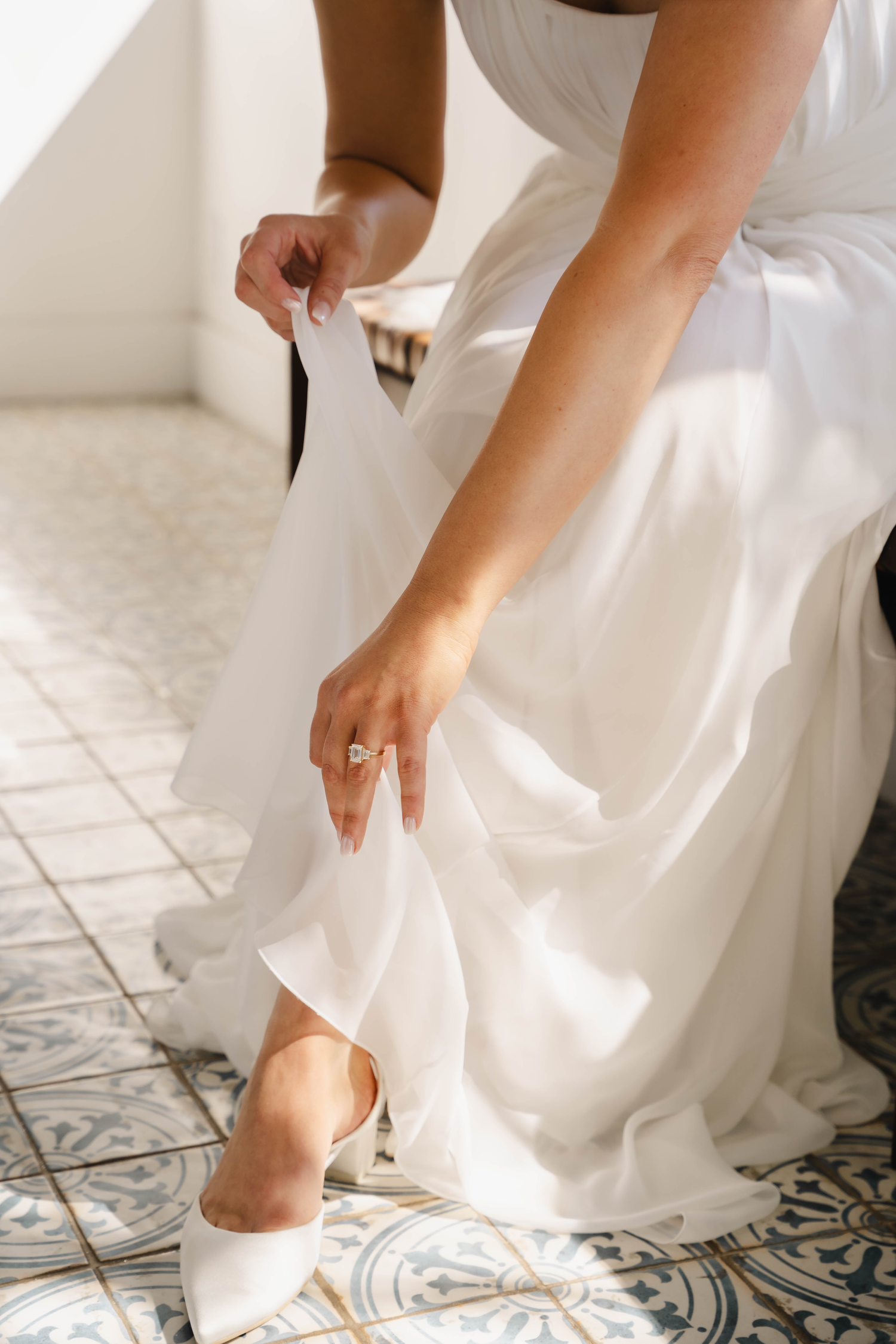 Bride in white wedding dress adjusting her white heels, showcasing elegant diamond engagement ring on tiled floor.