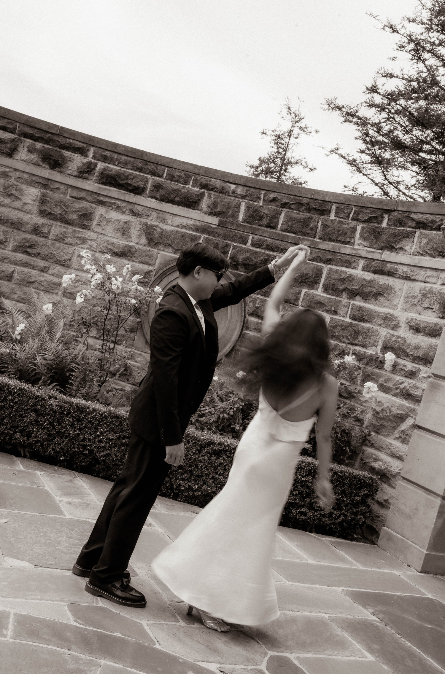 Black and white photo of a bride and groom dancing outdoors on stone patio with brick wall backdrop.