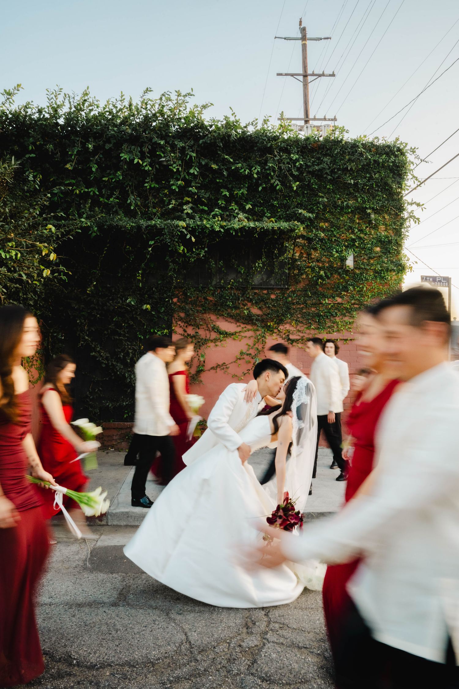 Bride and groom share a romantic dip kiss surrounded by wedding party in red dresses and white barong attire outdoors.