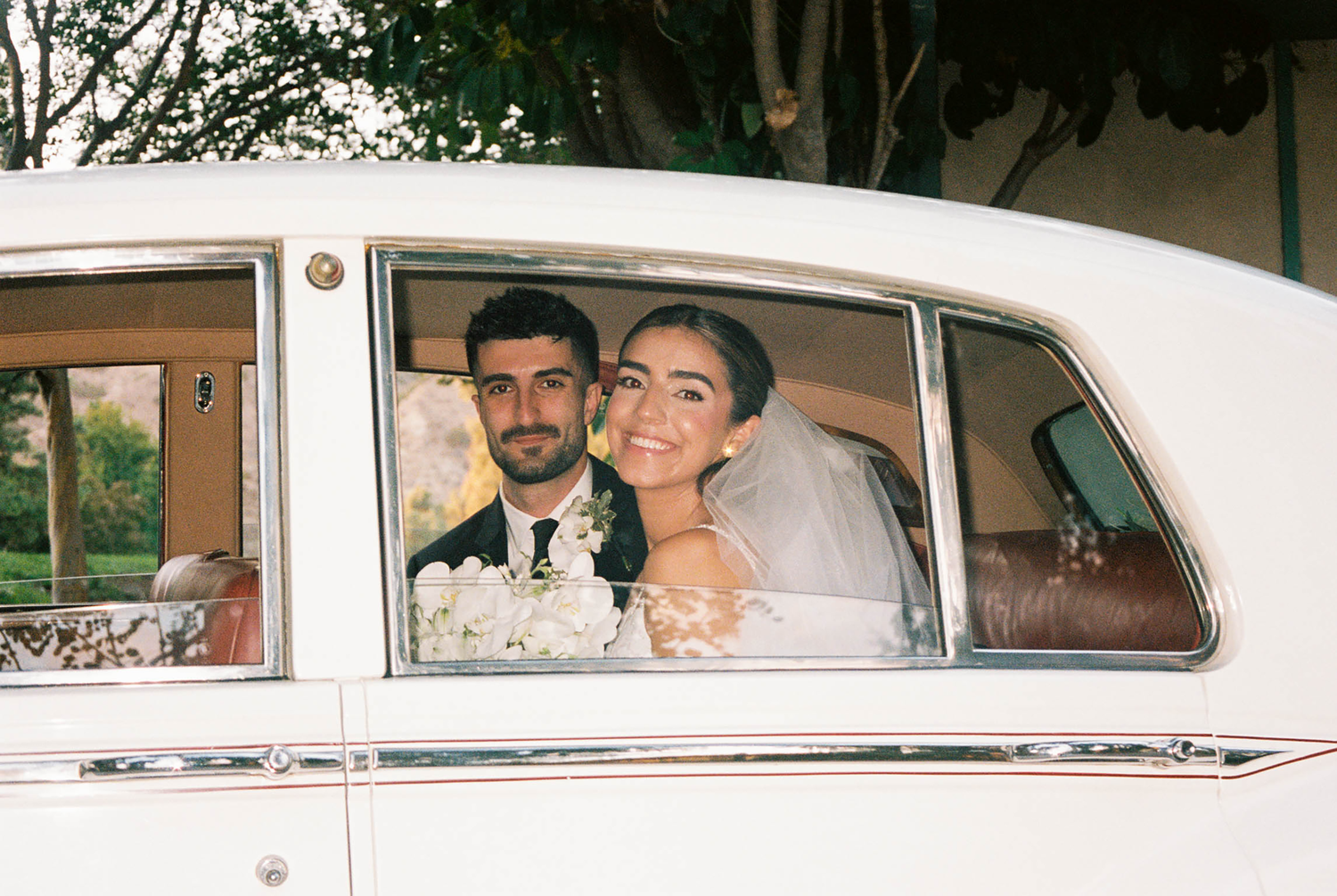 Smiling bride and groom seated inside vintage white classic car, bride holding white floral bouquet.