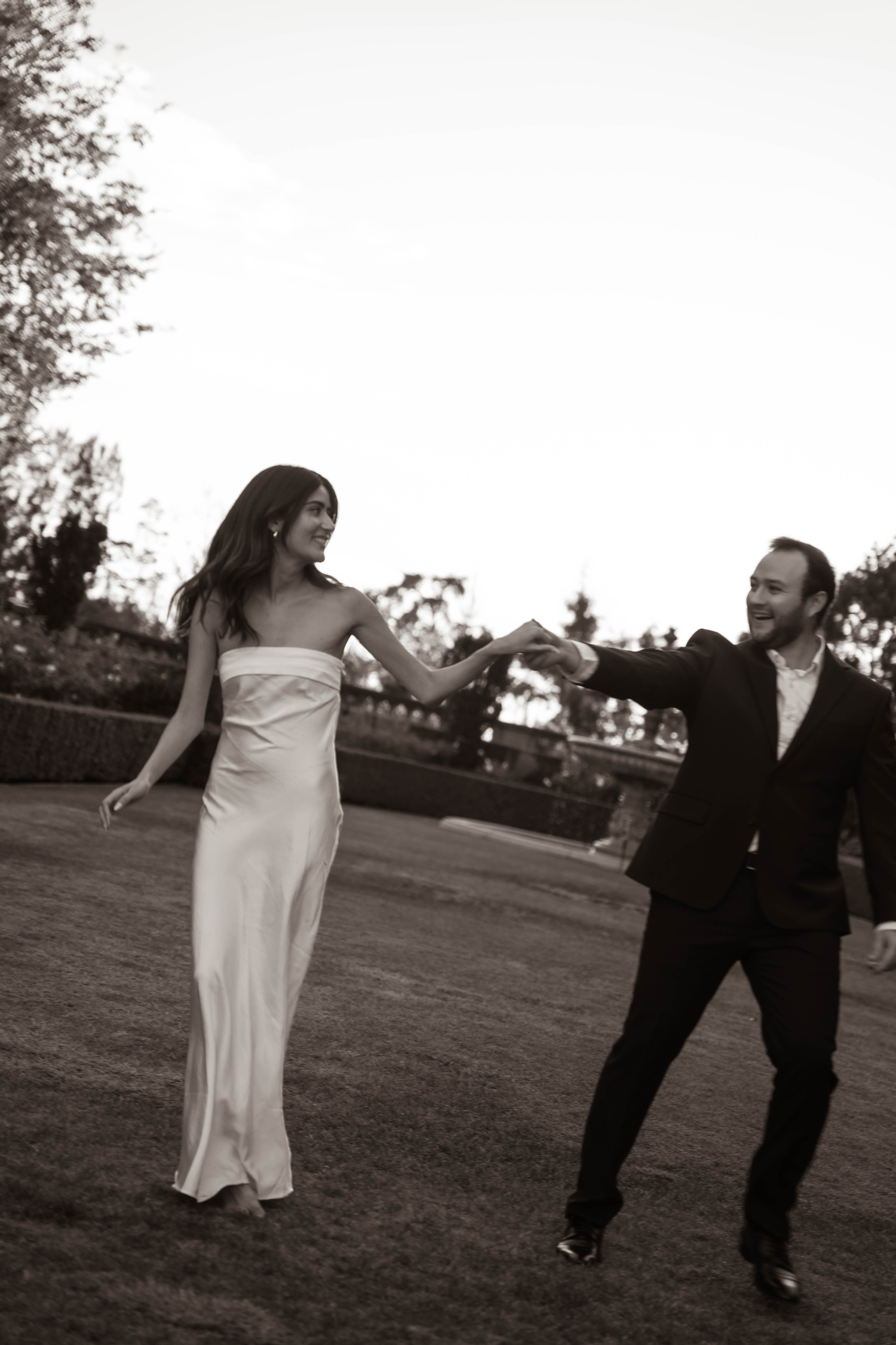 Black and white photo of bride in white gown and groom in suit dancing playfully on lawn.
