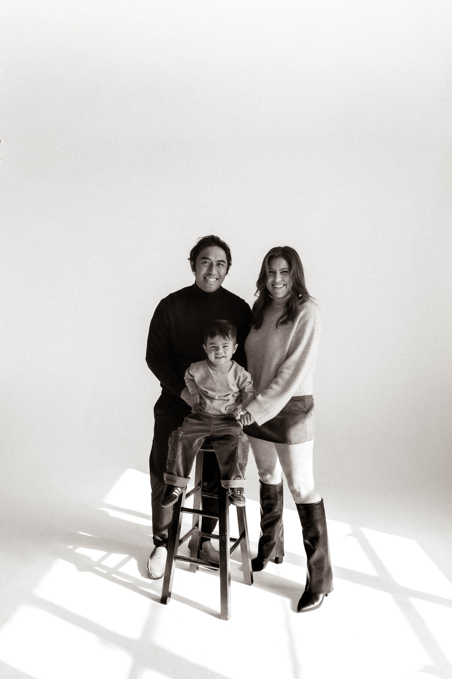 Black and white studio portrait of a family with two parents and young child seated on a stool.