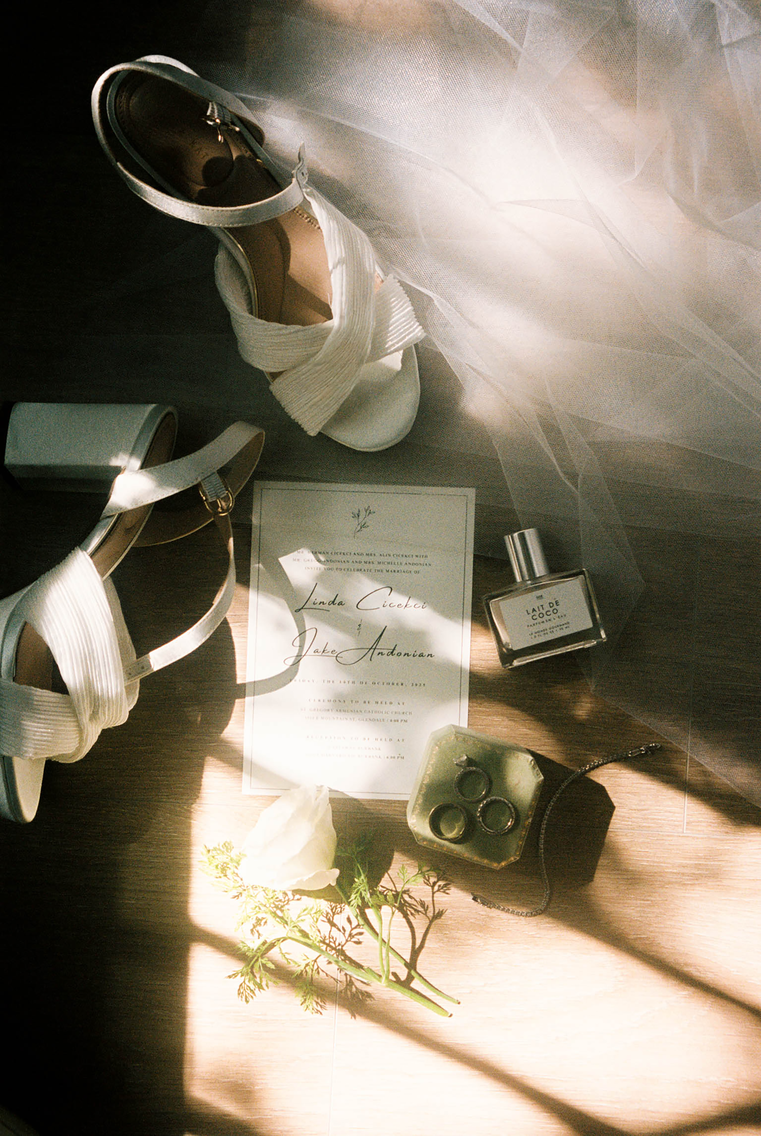 Flat lay of wedding details including white heels, invitation, perfume bottle, rings, and bridal veil in sunlight.
