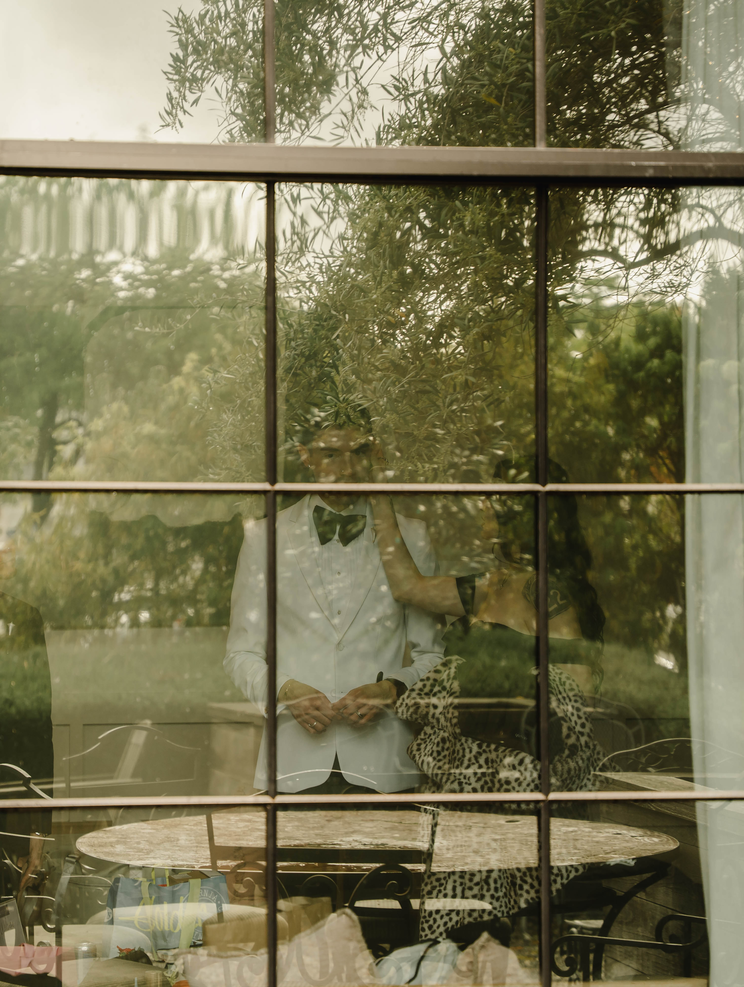 A person stands behind a multi-pane window surrounded by lush green trees, reflected in the glass.