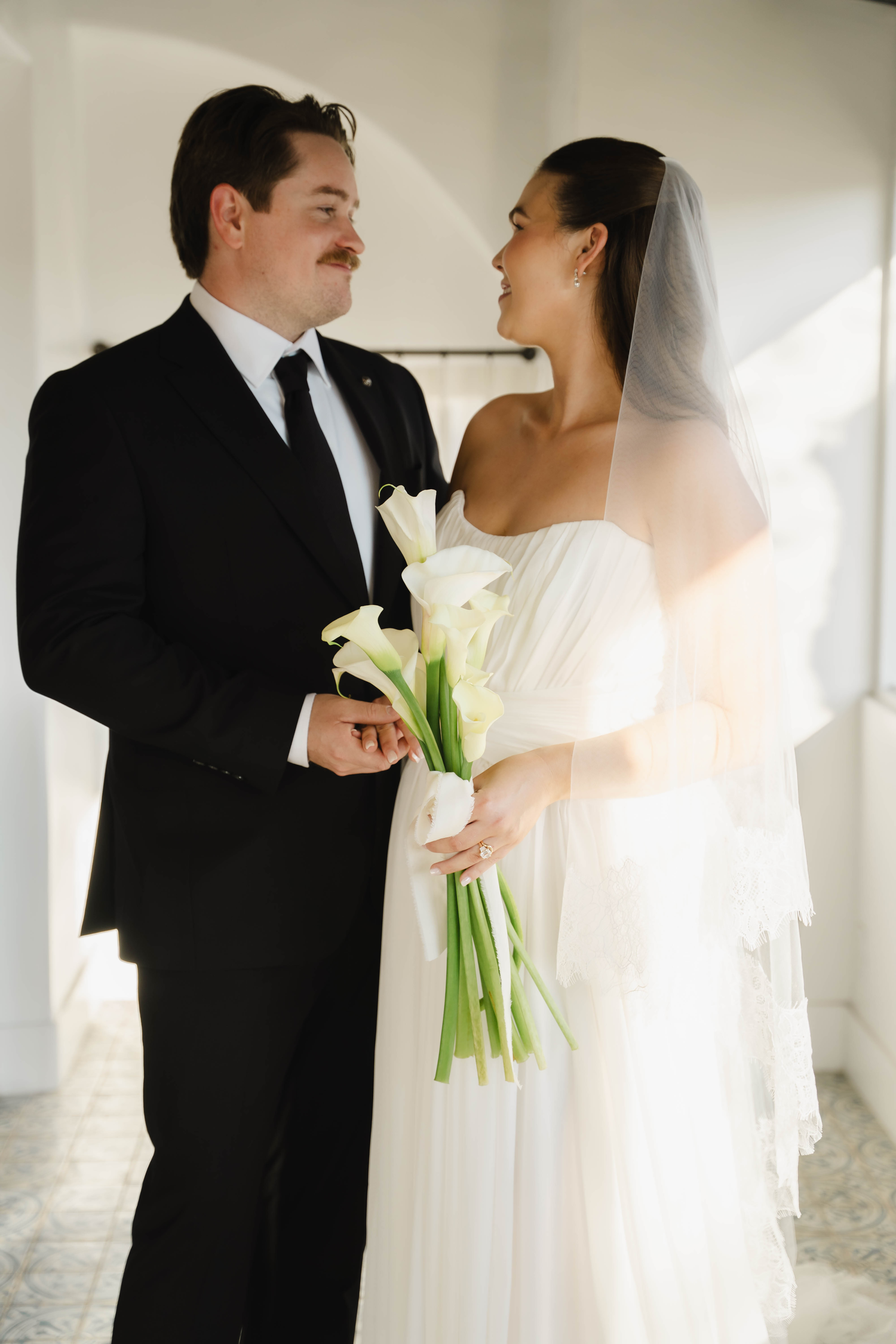 Bride in white gown holding calla lilies bouquet stands with groom in black suit, sharing a romantic gaze.