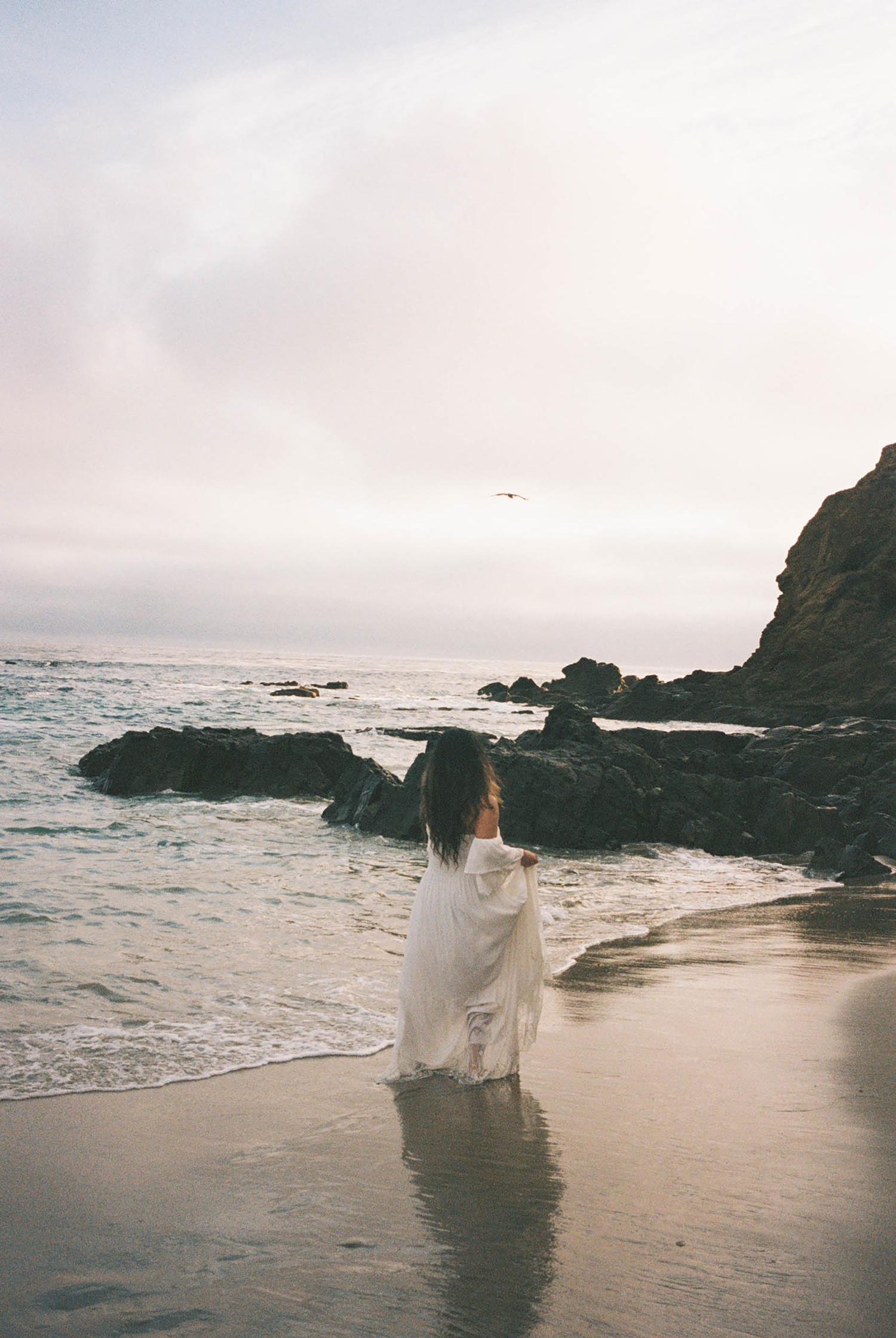 Pregnant woman in white lace dress standing on beach shoreline at sunset.