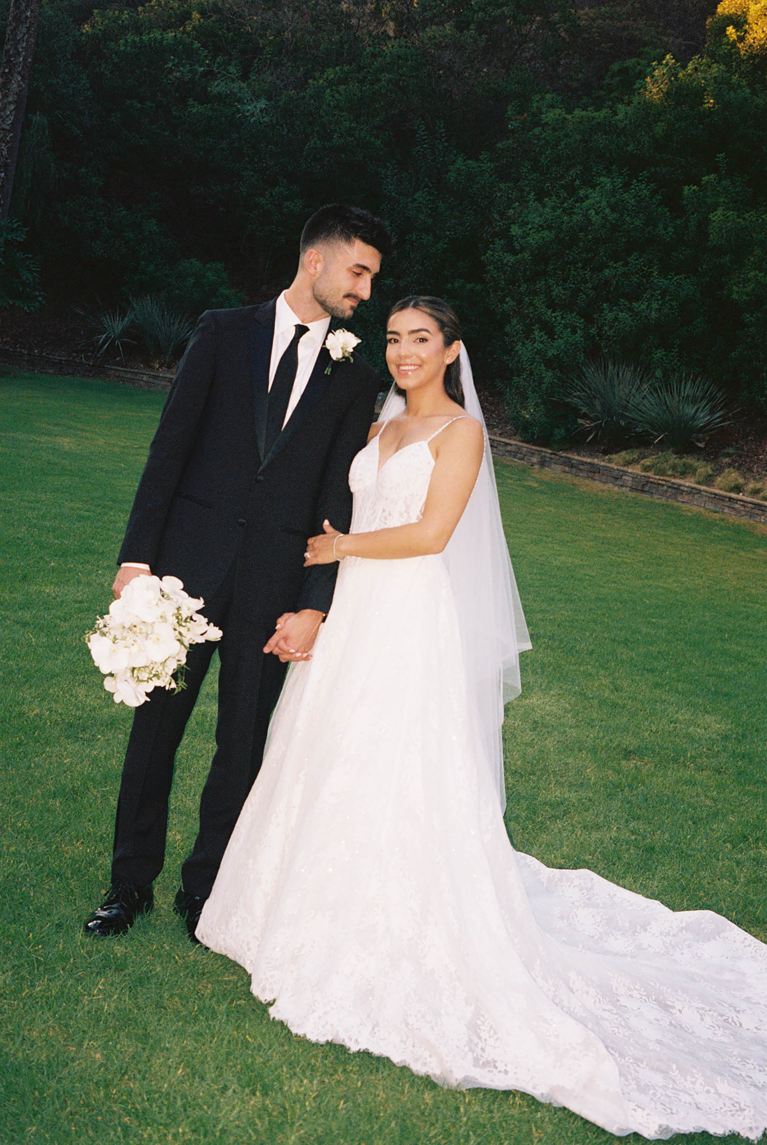 Wedding couple posing on green lawn, groom in black tuxedo holding bouquet, bride in lace gown with long veil.