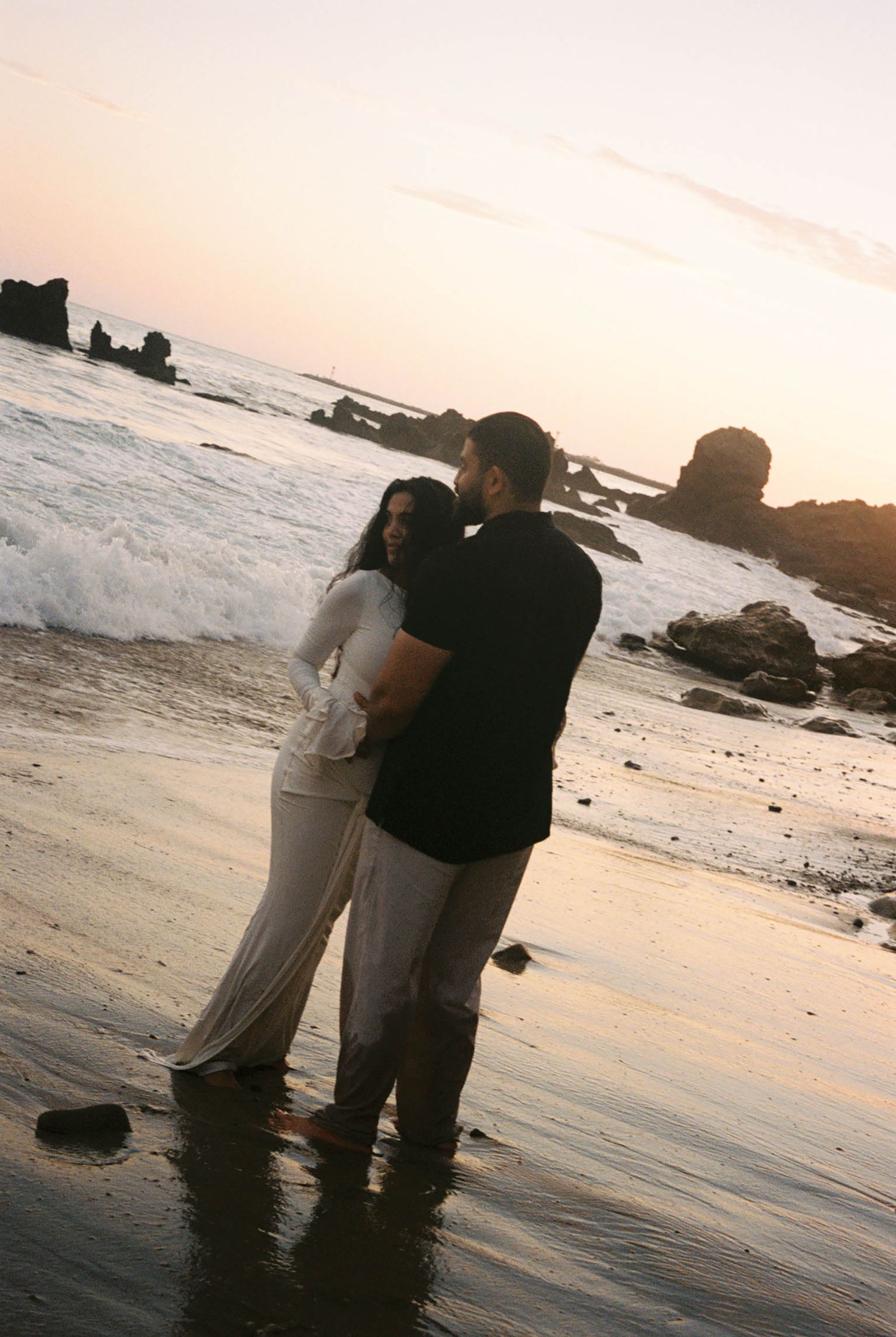A couple embraces romantically on a reflective beach at dusk with crashing waves behind them.