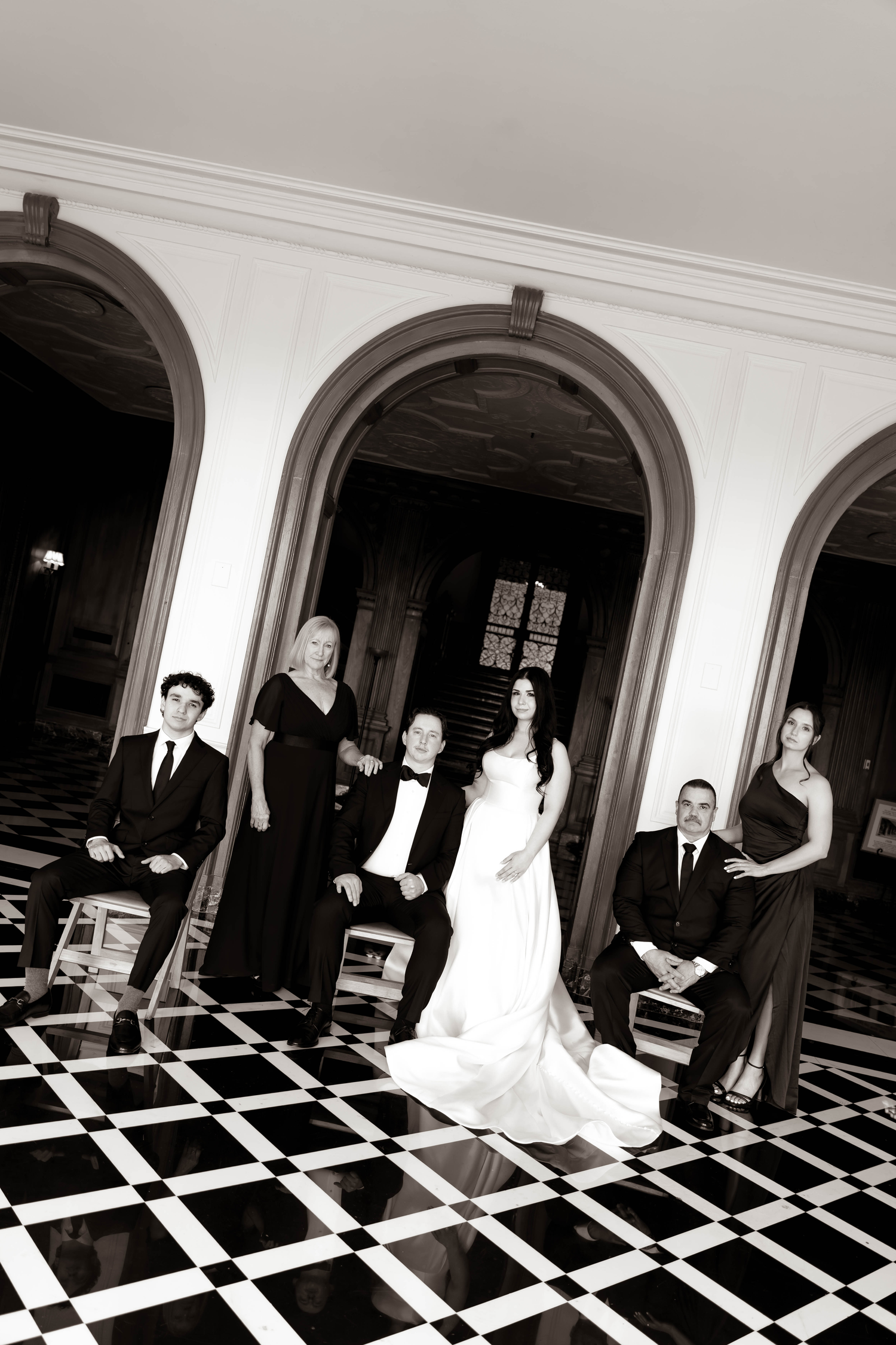 Black and white wedding photo of bride, groom, and family posing in elegant venue with checkered floors and arched doorways.