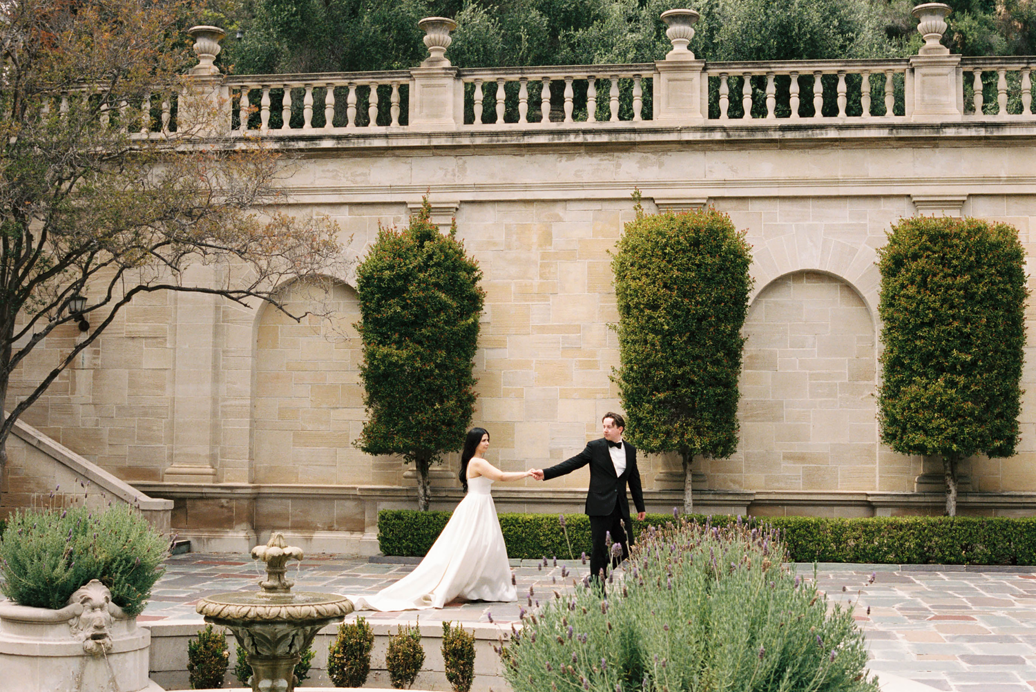 Bride and groom hold hands walking through elegant garden courtyard with stone arches and manicured hedges.