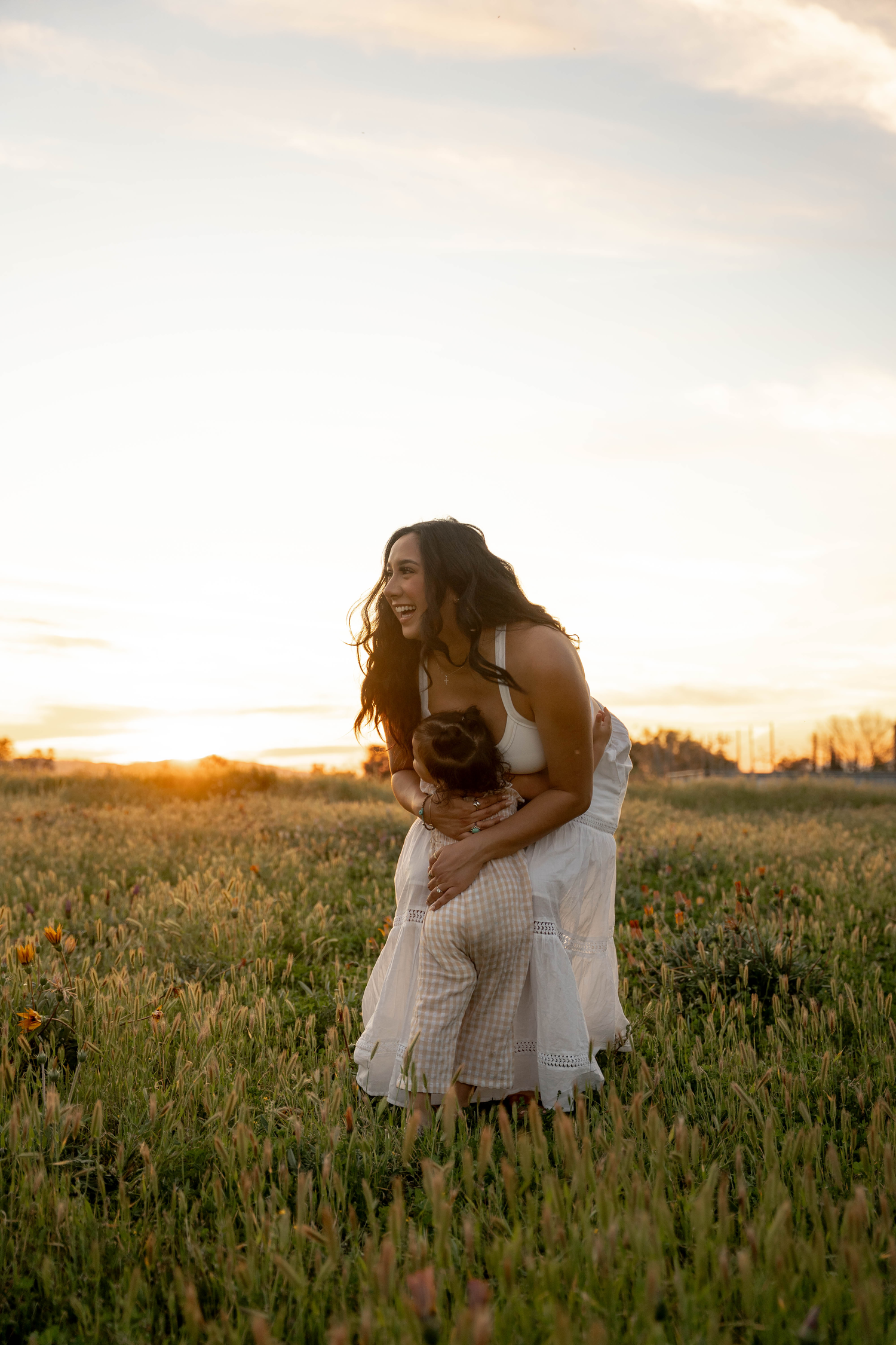A woman in a white dress laughs joyfully while embracing a child in a golden field at sunset.