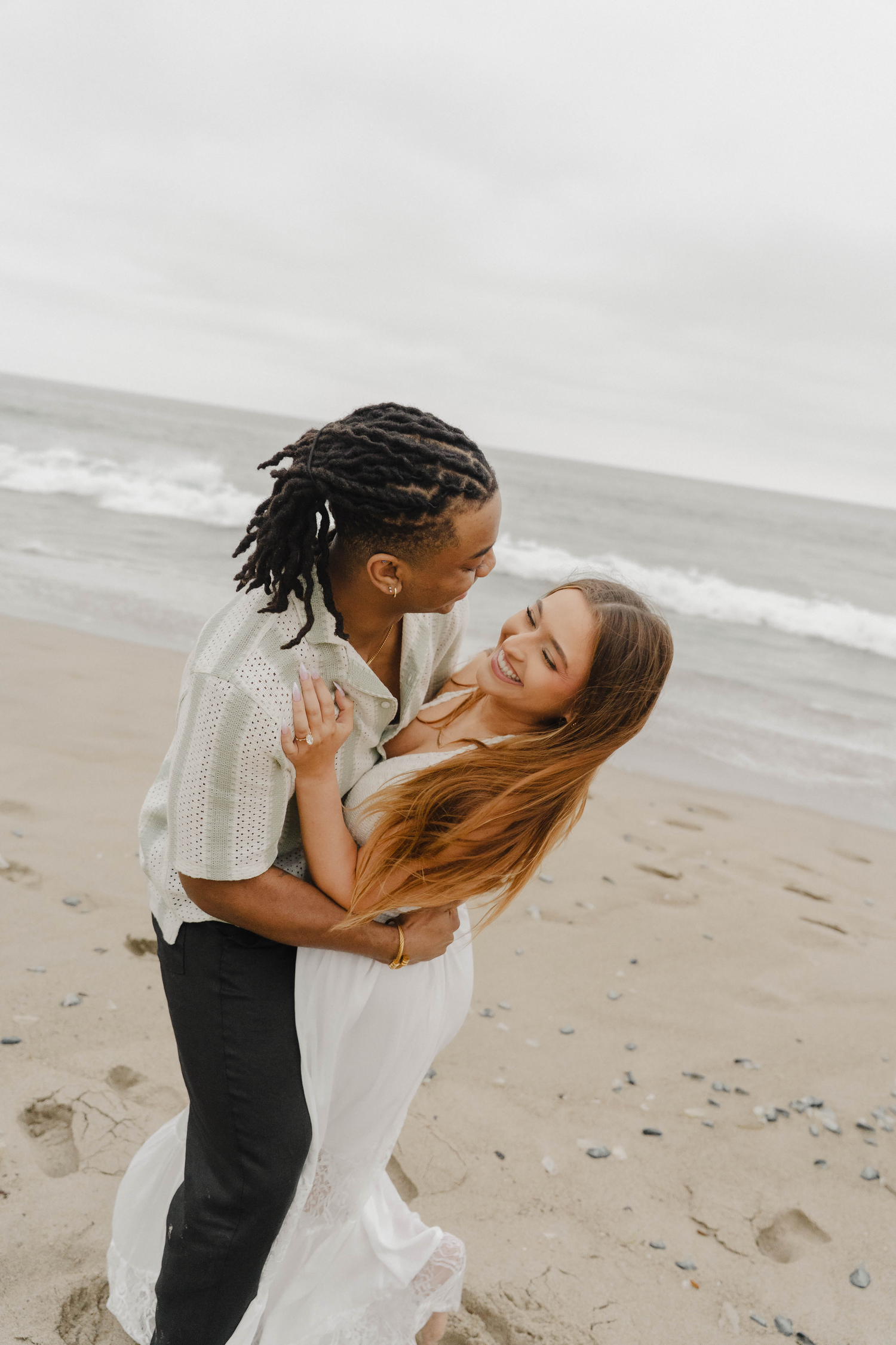 Couple embracing and smiling on a sandy beach with ocean waves, wearing casual white and light outfits.