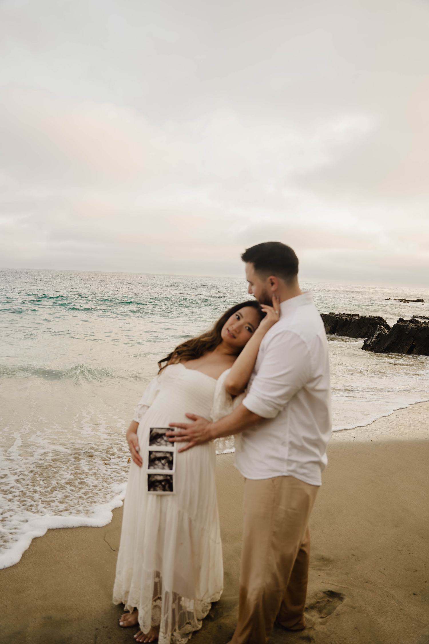 Pregnant couple embracing on beach at sunset, holding ultrasound photos, maternity shoot.