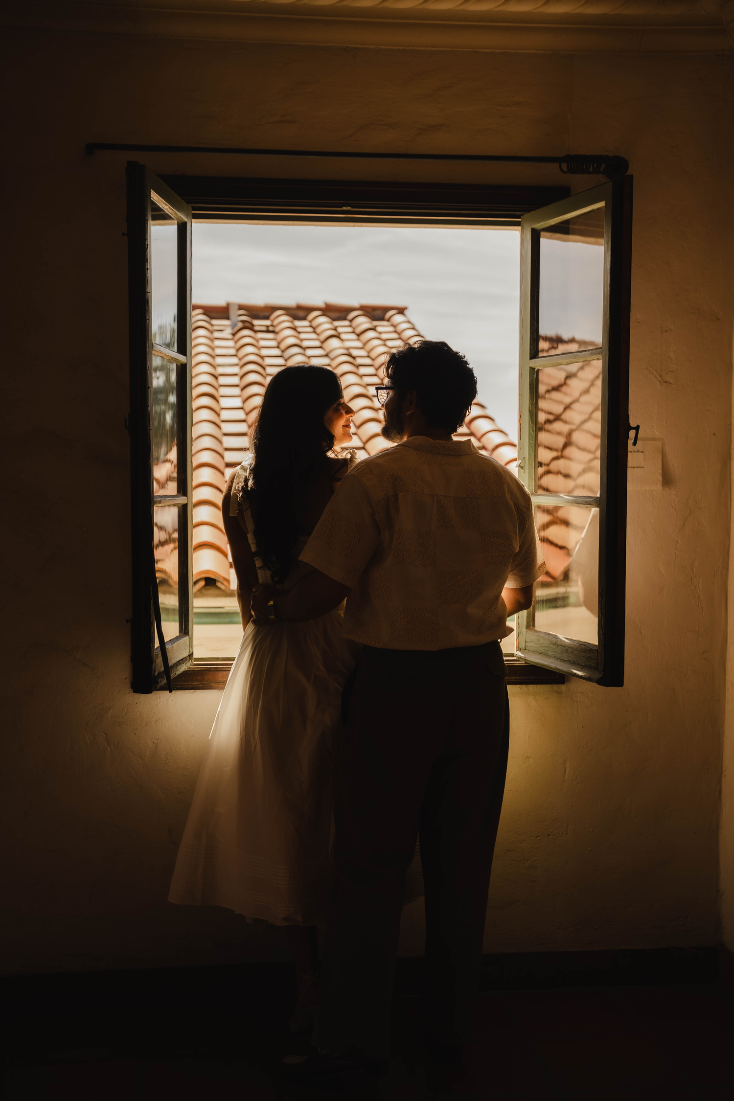 A couple stands silhouetted together at an open window overlooking terracotta rooftops at dusk.