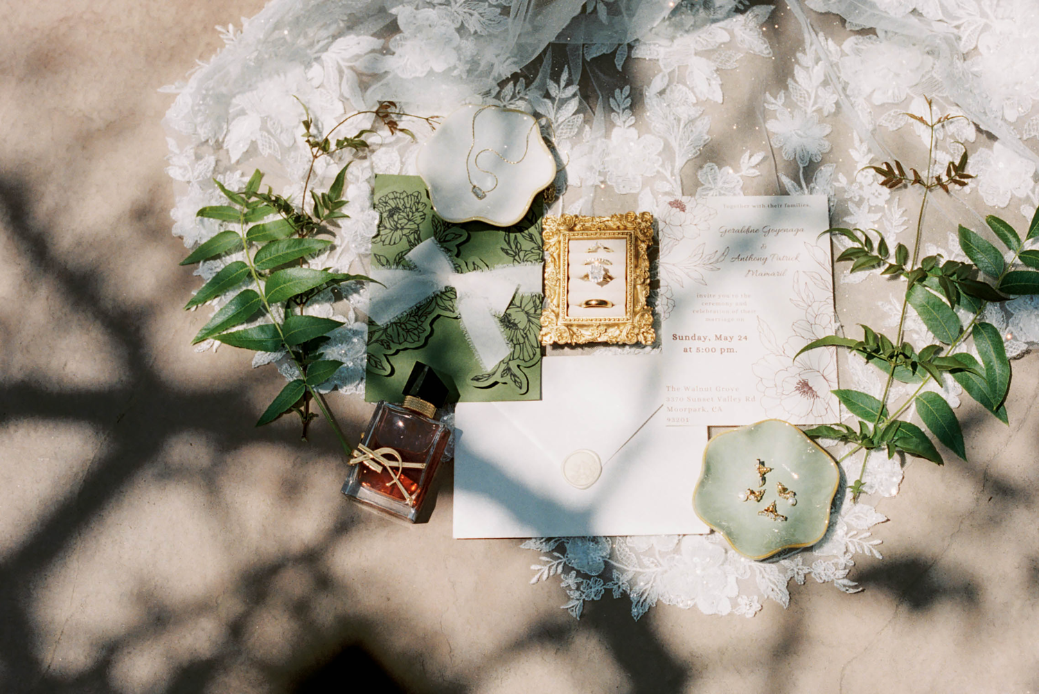 Elegant wedding flat lay with ivory invitations, gold frame, greenery, and floral accents on a soft white backdrop.