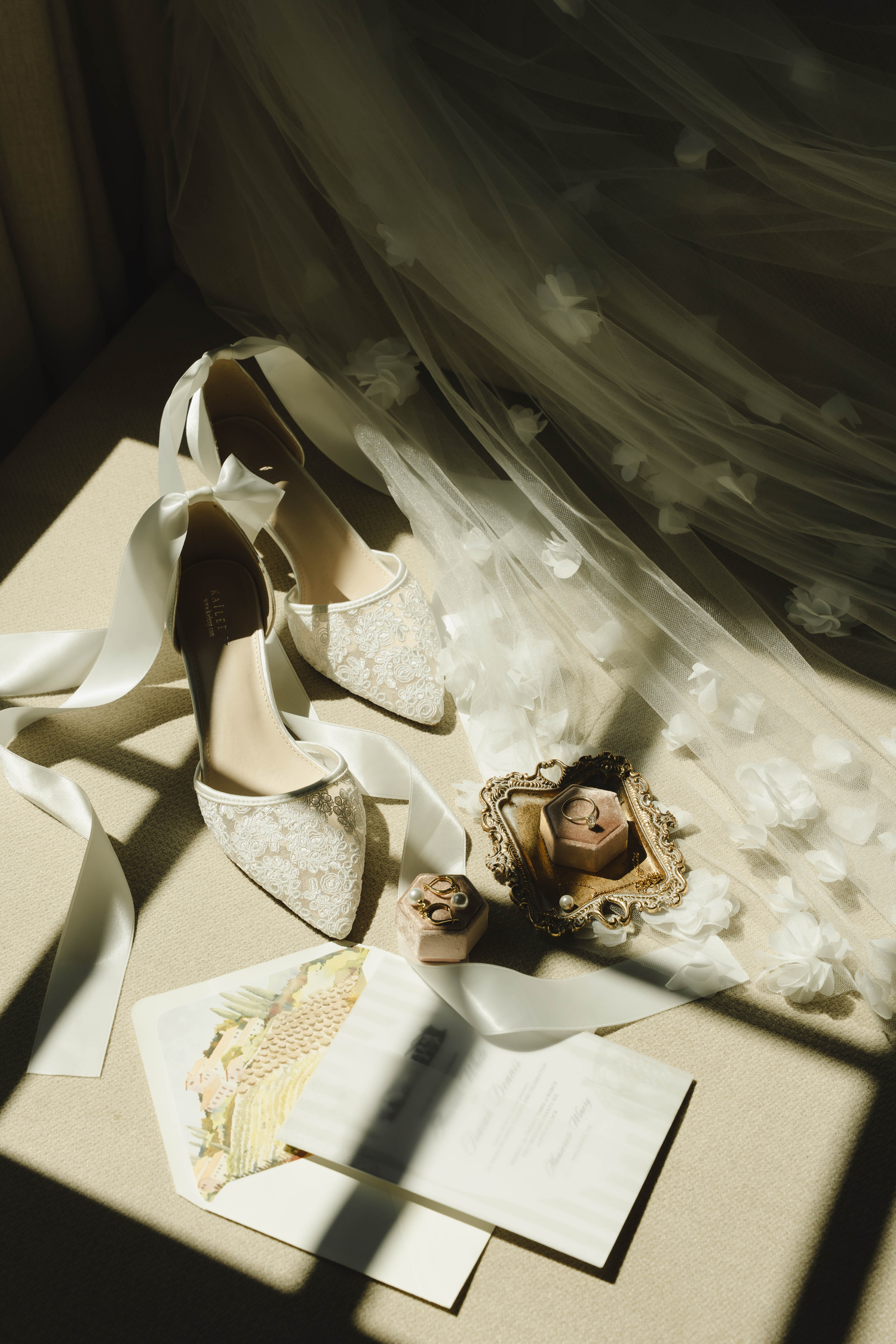 Elegant bridal flat lay with lace heels, white veil, wedding rings, and invitation cards in soft natural light.