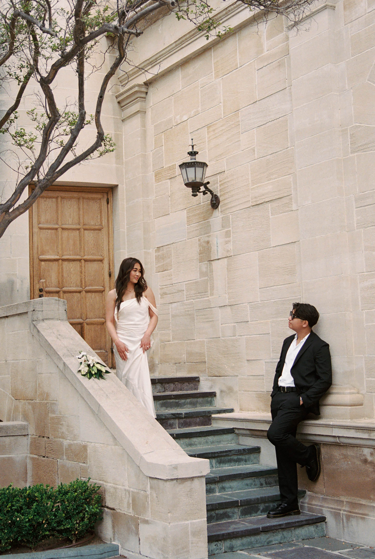 Bride in white gown and groom in black suit pose on stone steps outside an elegant building.