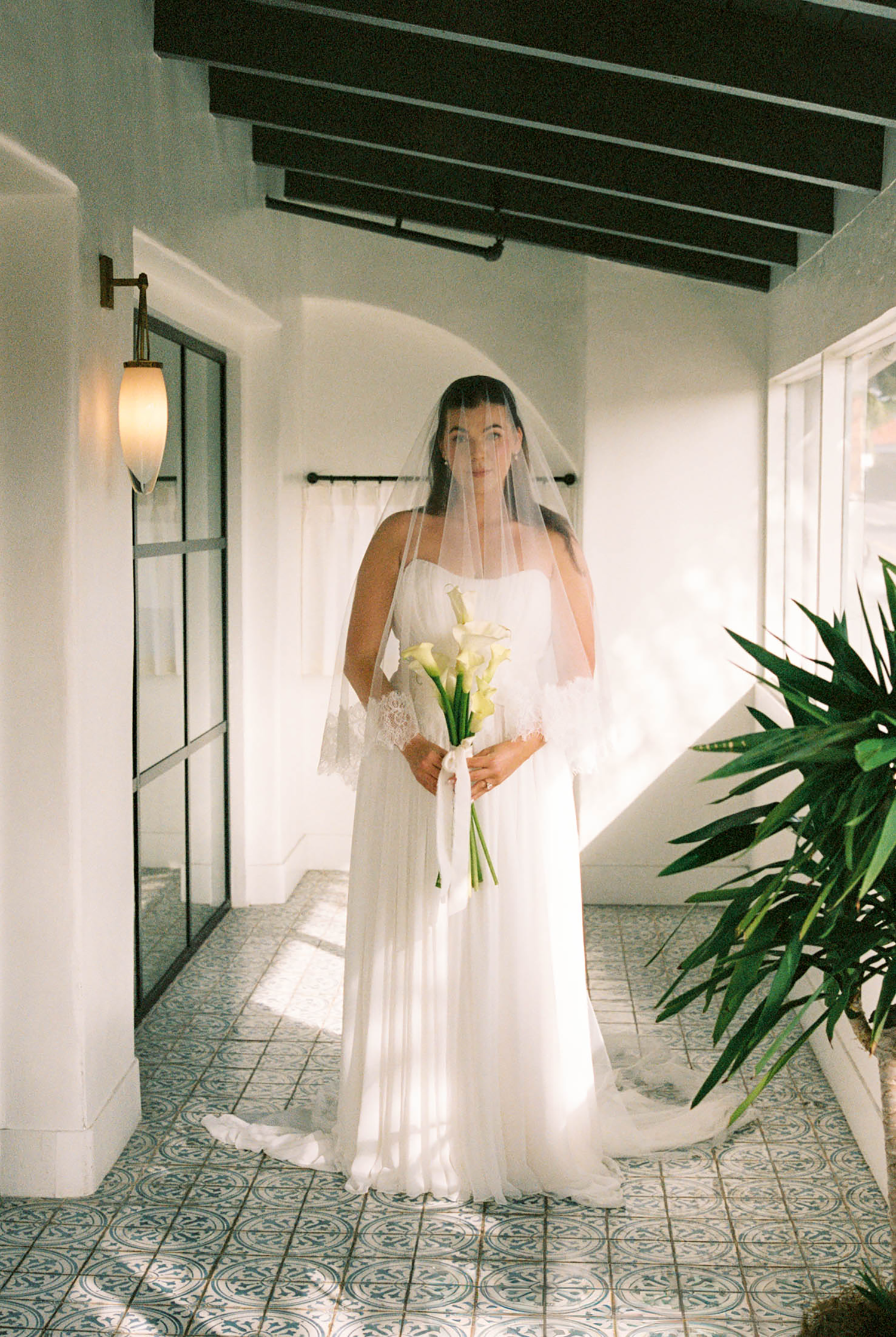 Bride in white flowing gown and veil holding calla lily bouquet standing in sunlit Spanish-style venue hallway.
